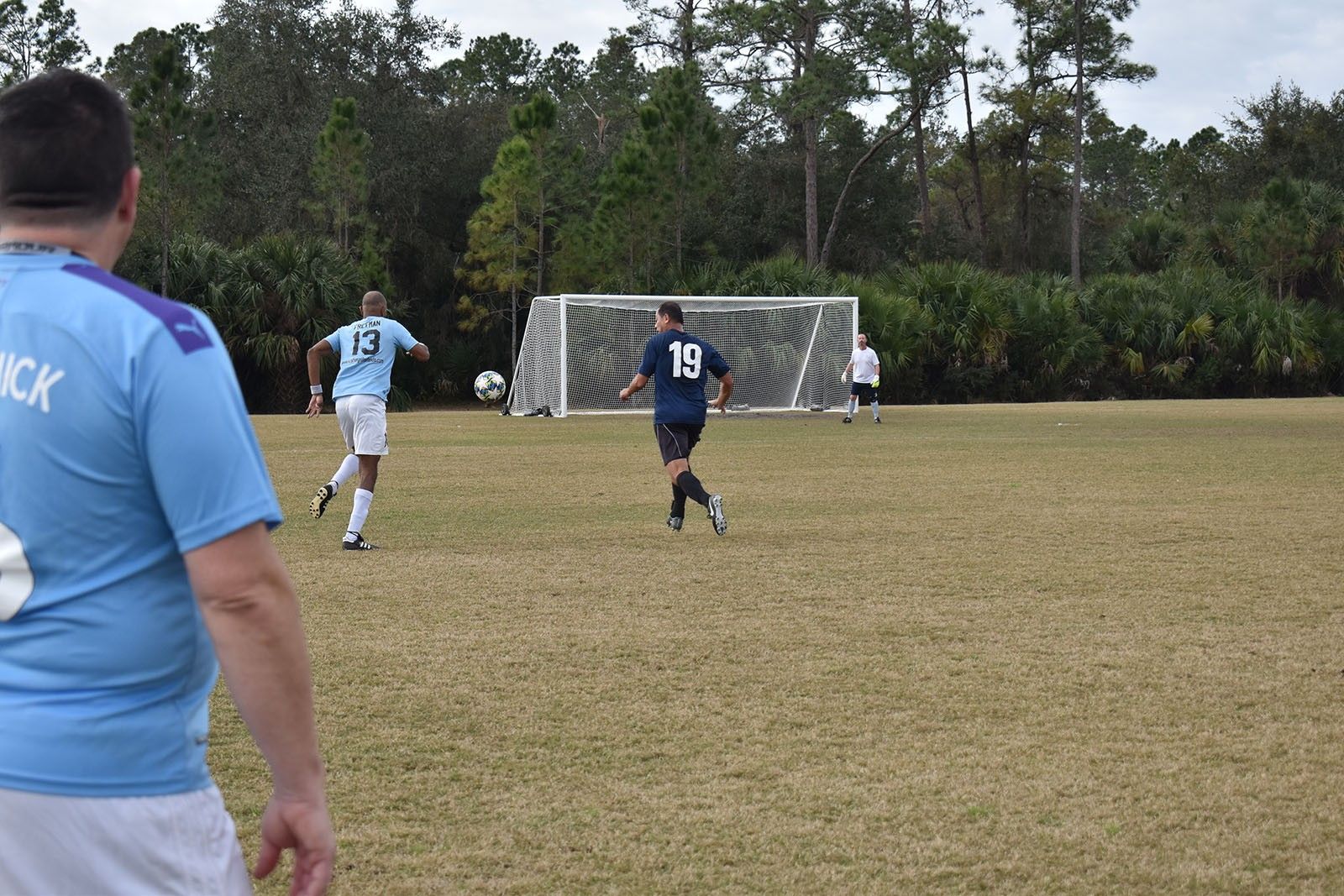 Soccer players run toward the goal on a grassy field, with a goalkeeper near the net.