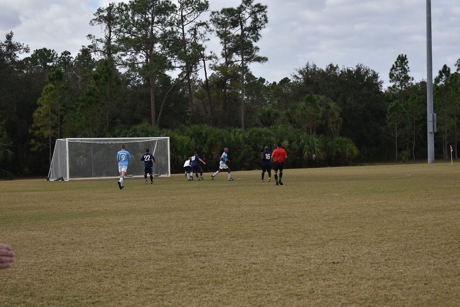 Soccer players near a goal on a grassy field, with one in red running toward the action.