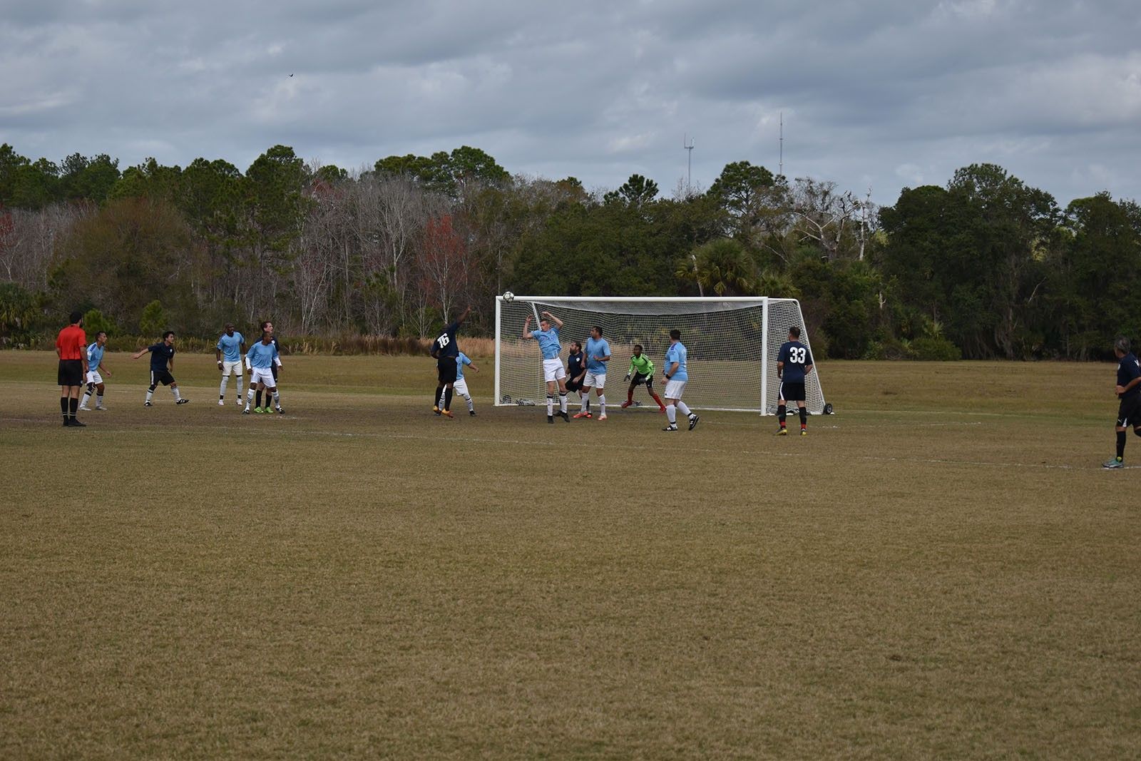 Soccer players gather near the goal on a muddy field under an overcast sky.