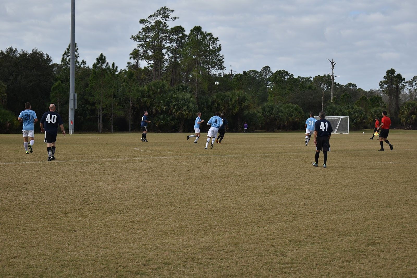 Soccer players spread across a grassy field during a match, with trees and utility poles in the background