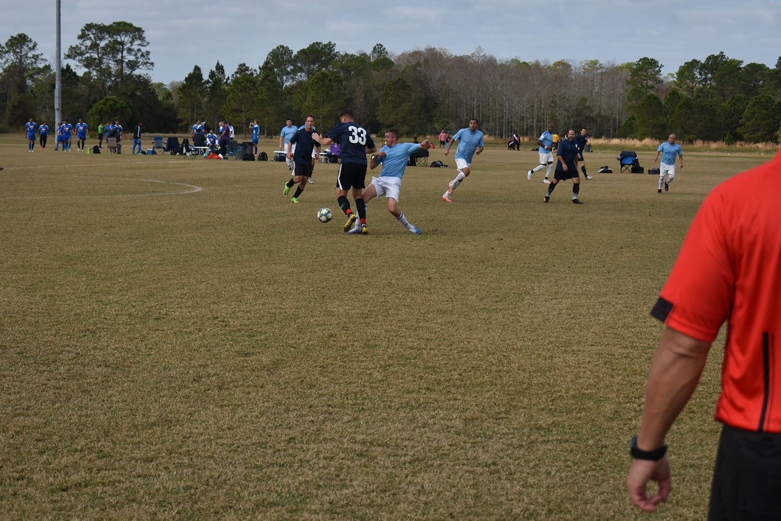 Soccer players on a grassy field, with one player in red in the foreground and spectators in the distance