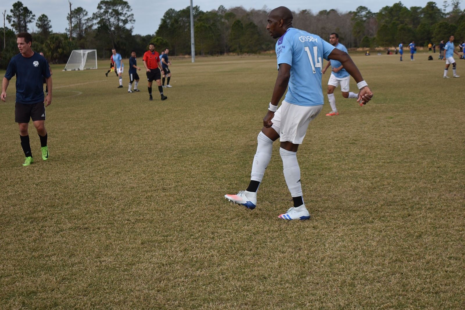 Soccer players on a grassy field, with one in a light blue jersey walking in the foreground.