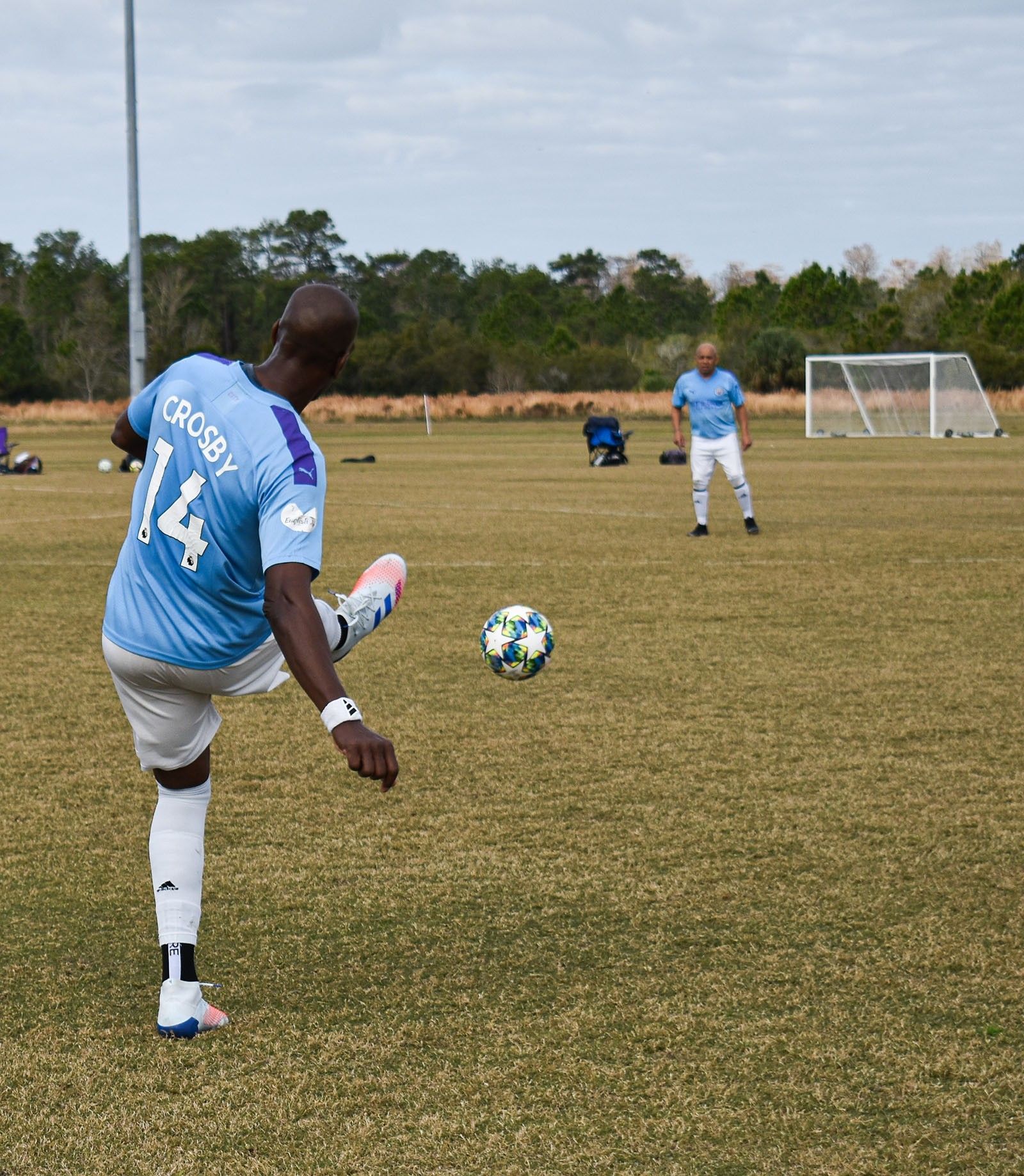 Soccer player in blue jersey kicks a ball on a grassy field toward a teammate near the goal.