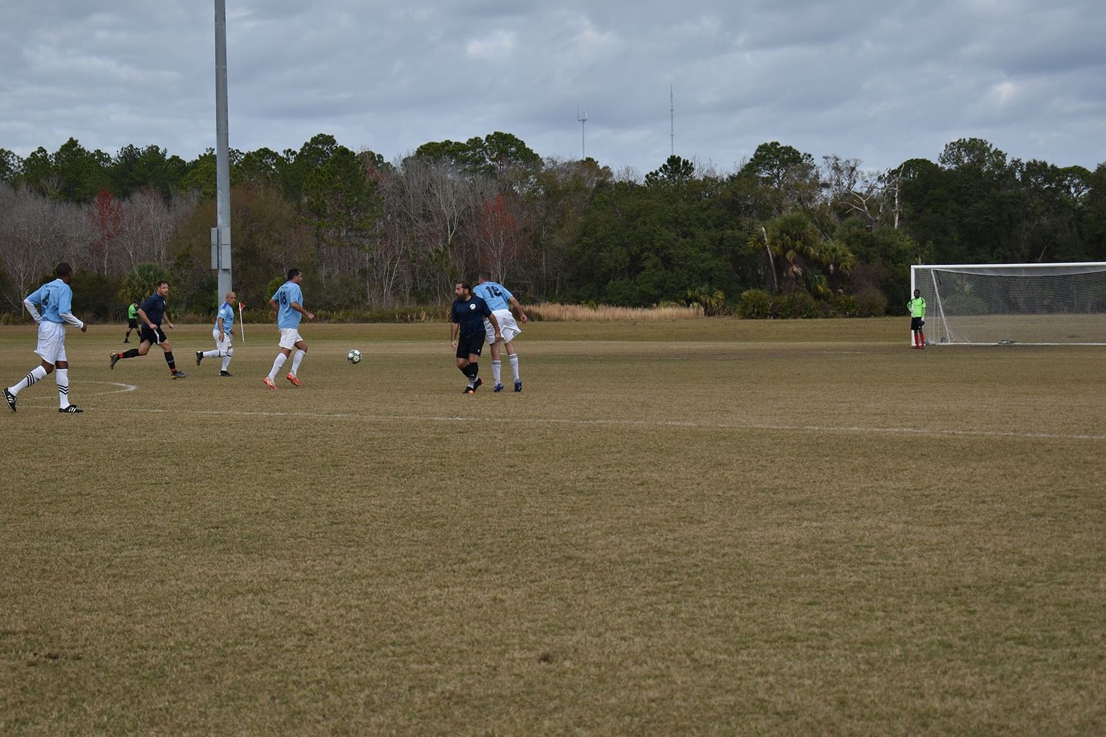 Soccer players in blue and black compete on a field near the goal under cloudy skies