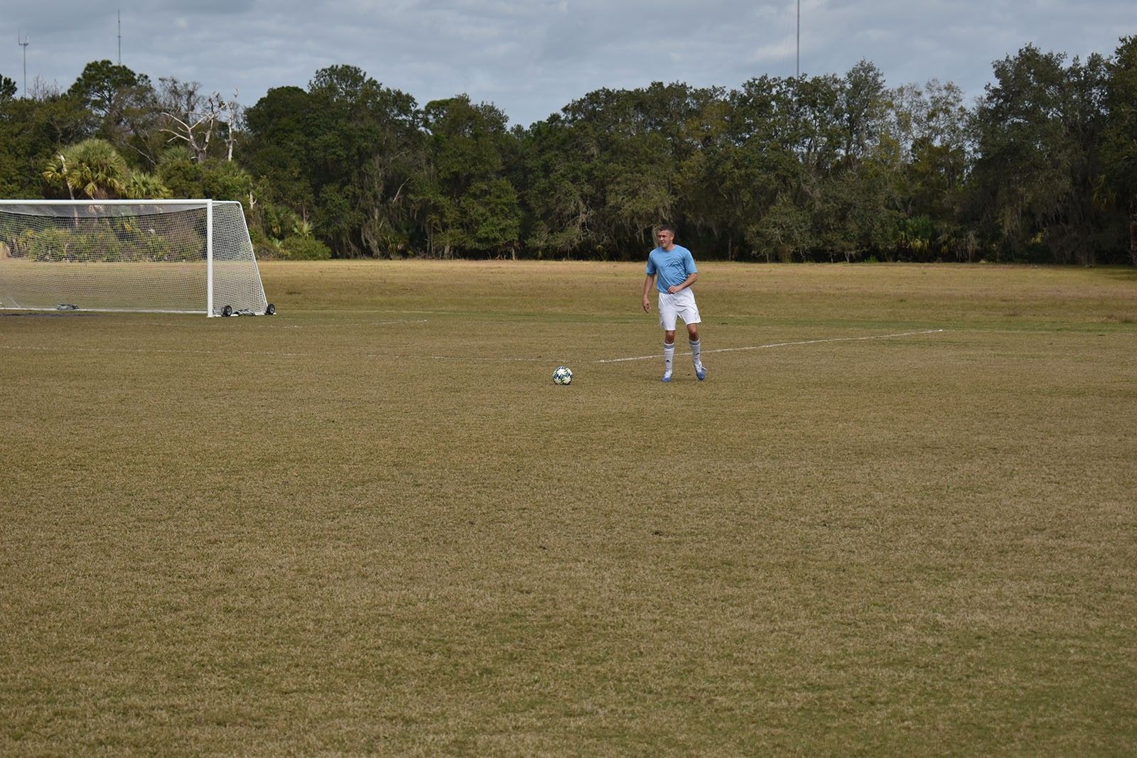 Soccer field with a lone player in blue standing near a ball, goal visible on the left.