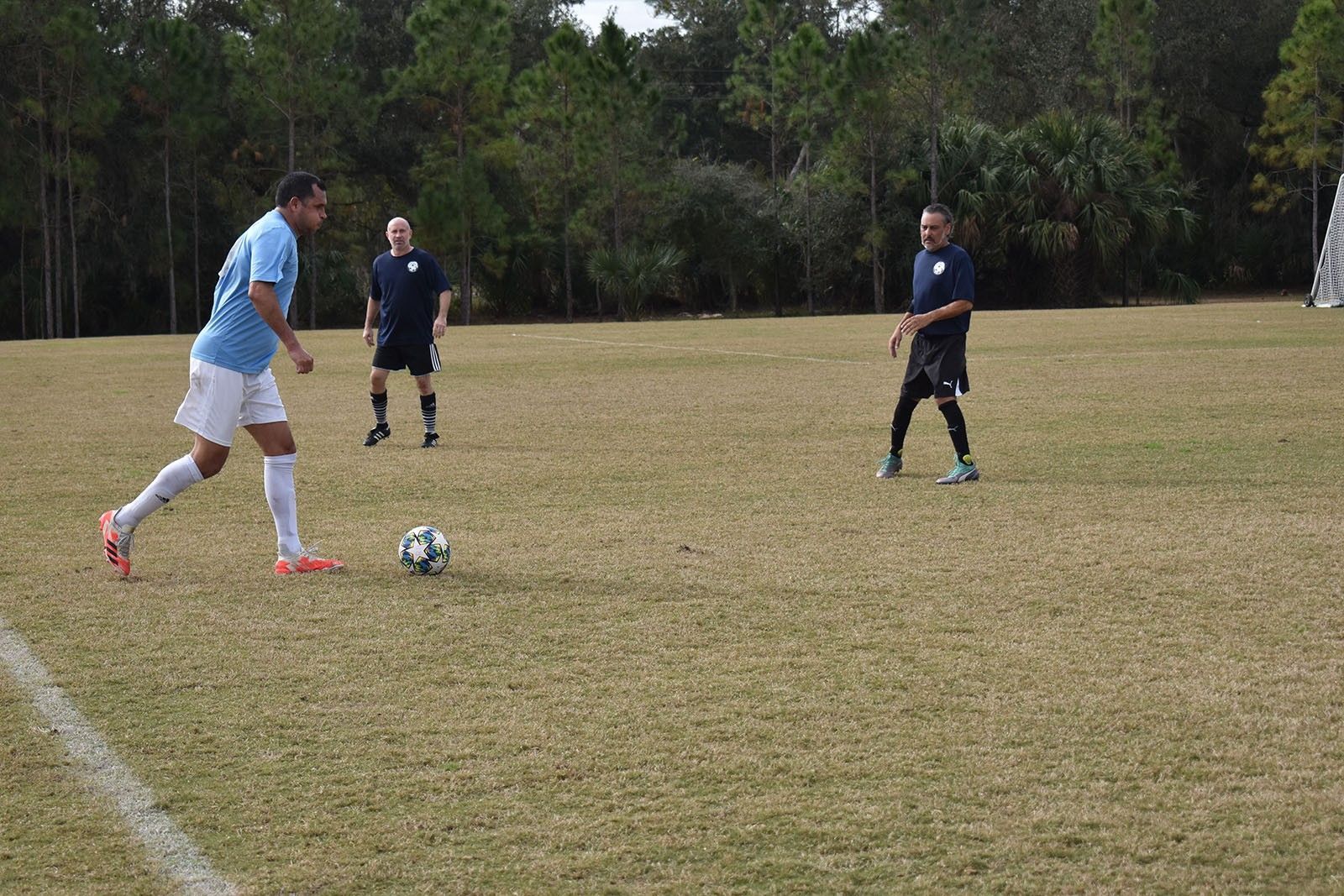 Soccer player in light blue prepares to kick a ball on a grassy field with two defenders nearby