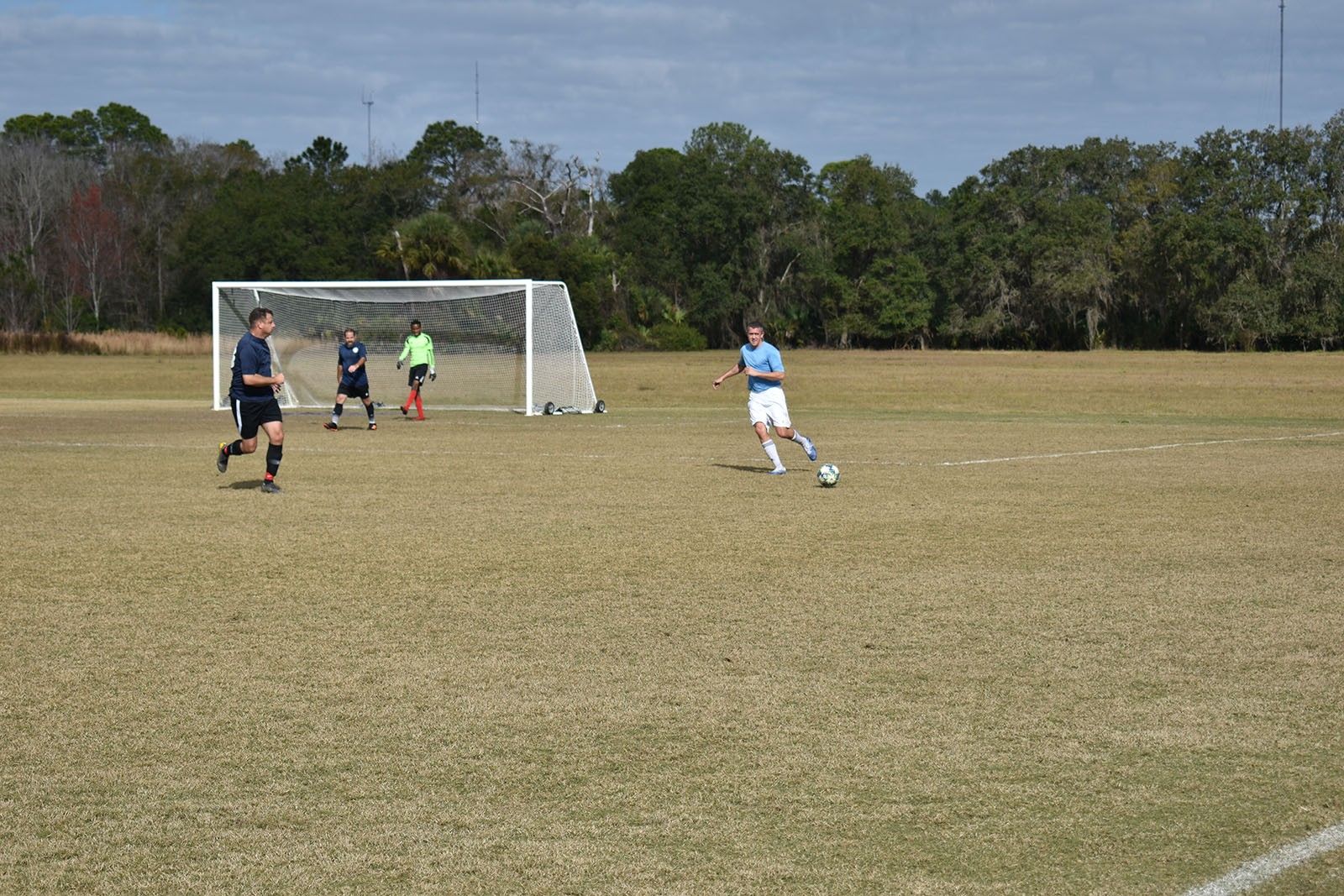 Soccer players on a grassy field near a goal, with one in white running toward the ball.