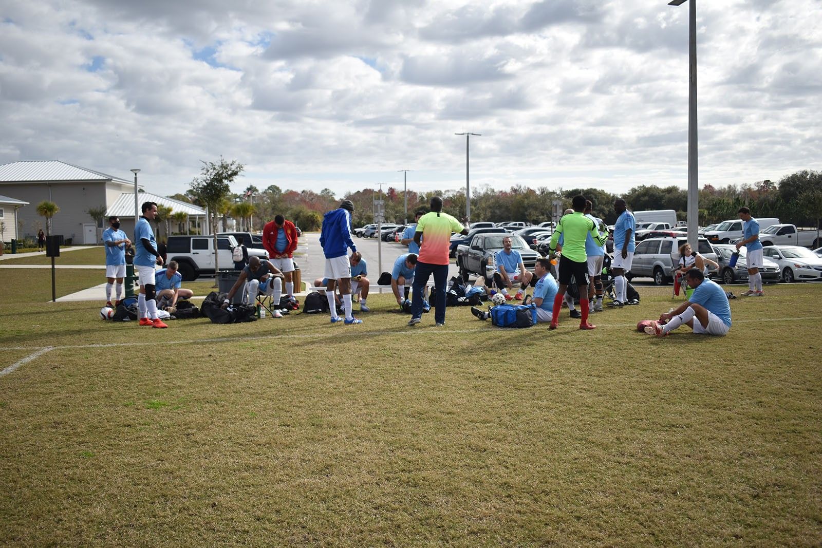 Youth soccer game with players and coaches gathered on a grassy field near parked cars