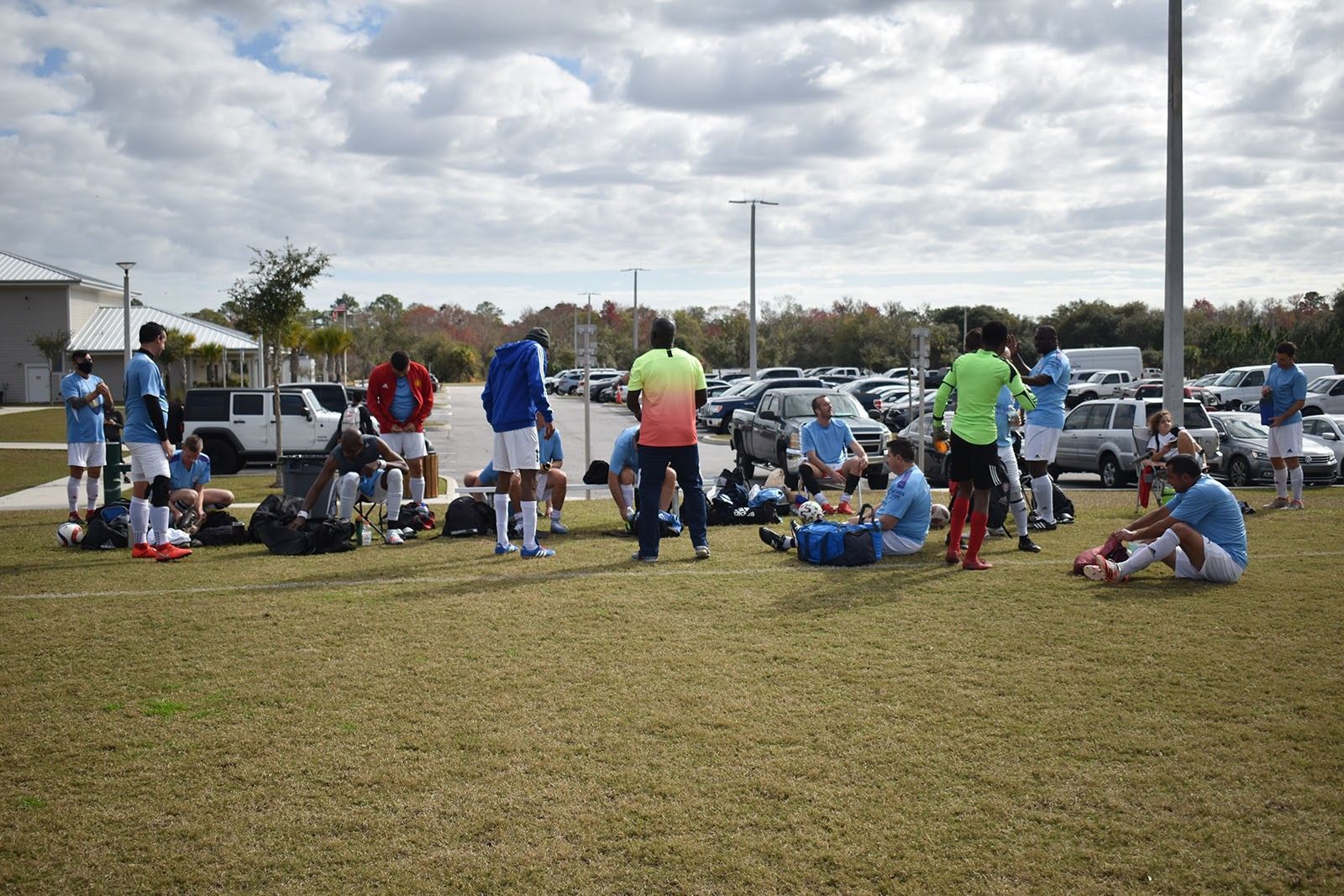 Soccer team resting on a grassy field near parked cars, with players and coaches gathered around equipment.