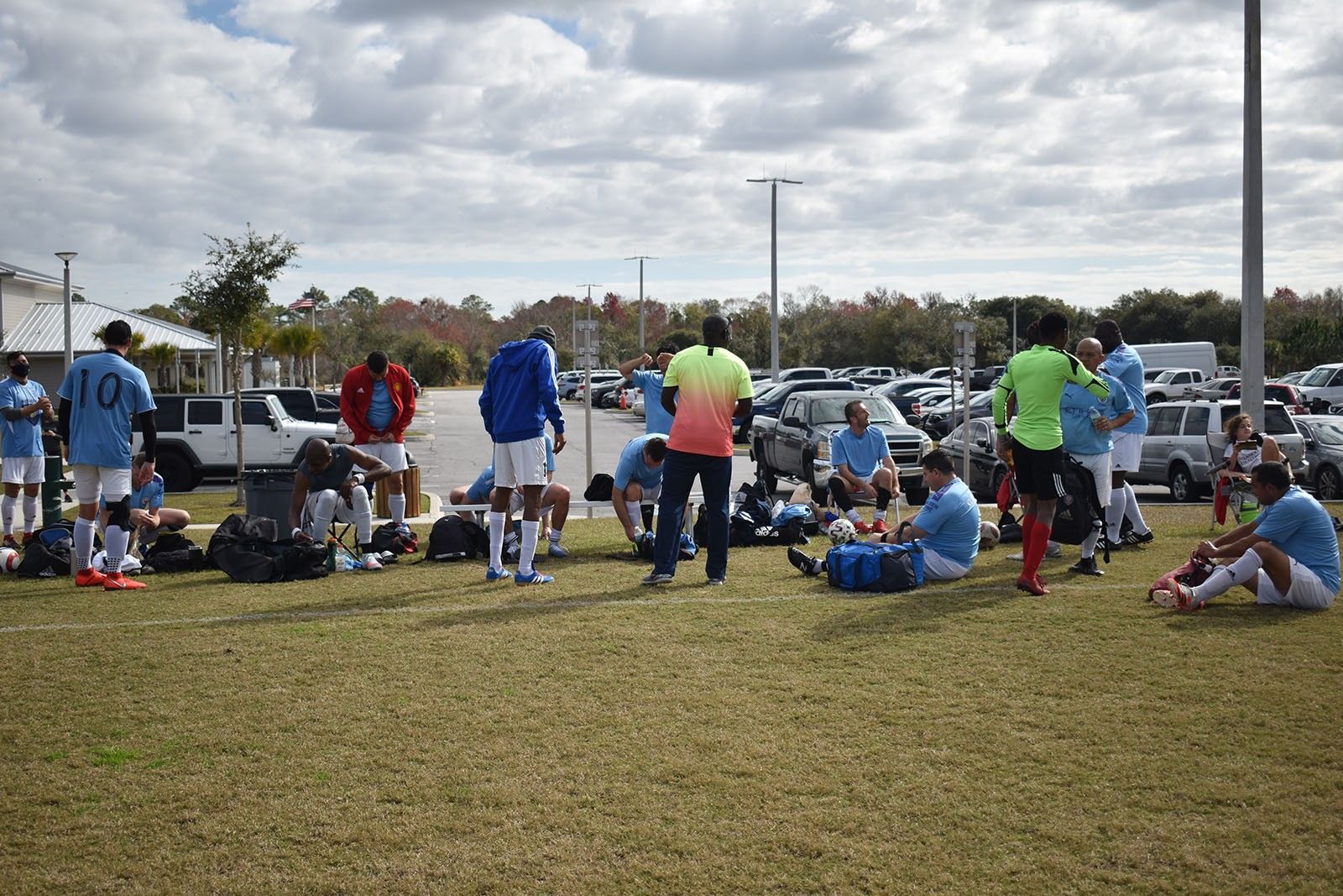 Soccer players and spectators gathered on a grassy field near parked cars and portable benches.