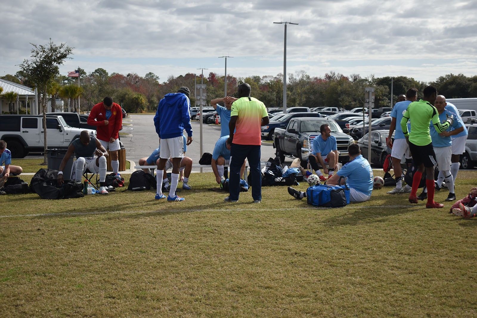 People in sports gear gathering on a grassy field with bags and coolers near parked cars