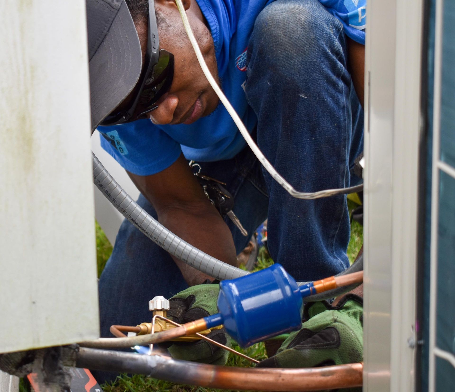 Worker in blue shirt and goggles repairs copper pipes inside a cramped utility space.