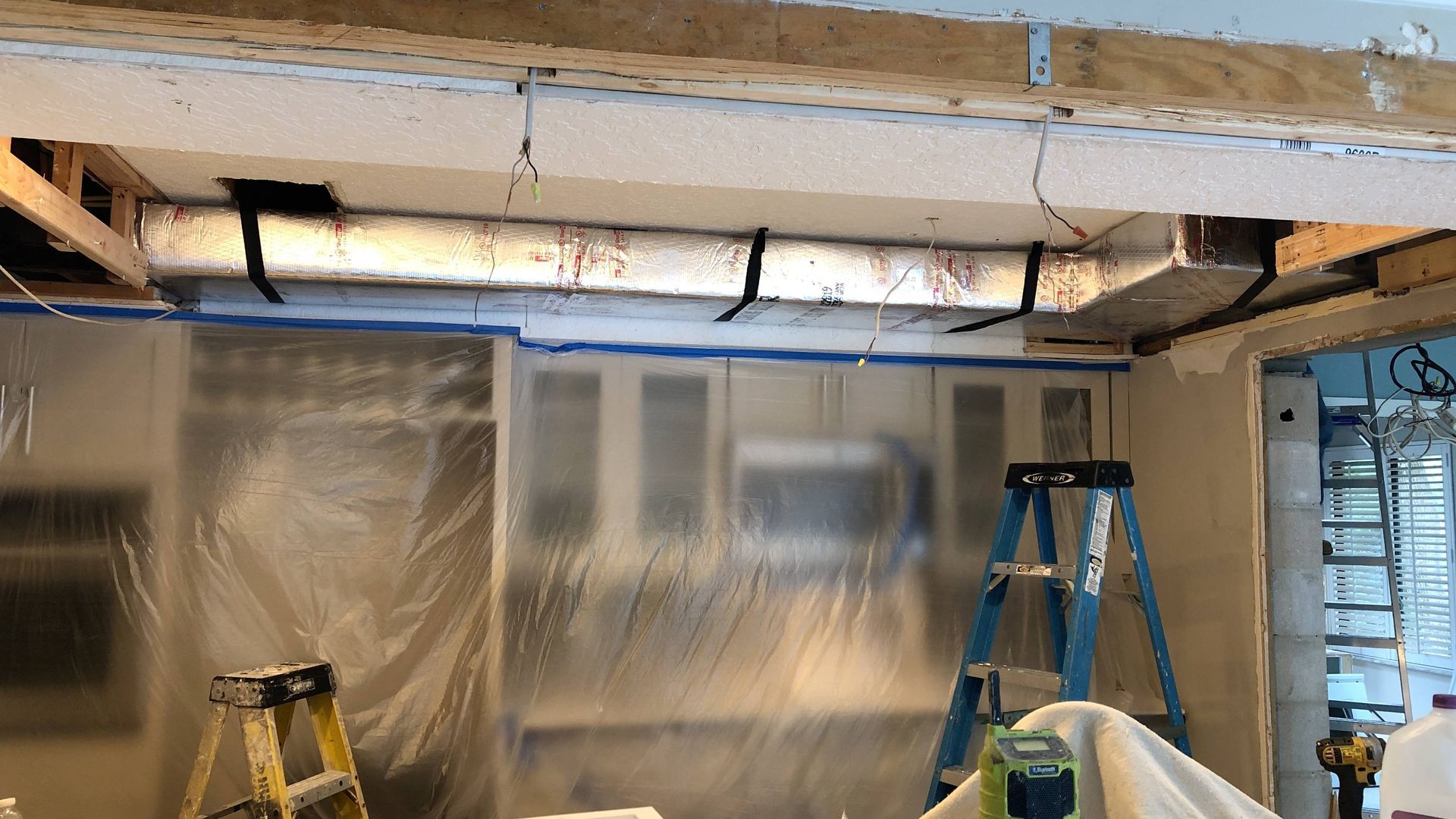 Partially renovated room with exposed ceiling, plastic sheeting, and a blue ladder over a covered wall opening