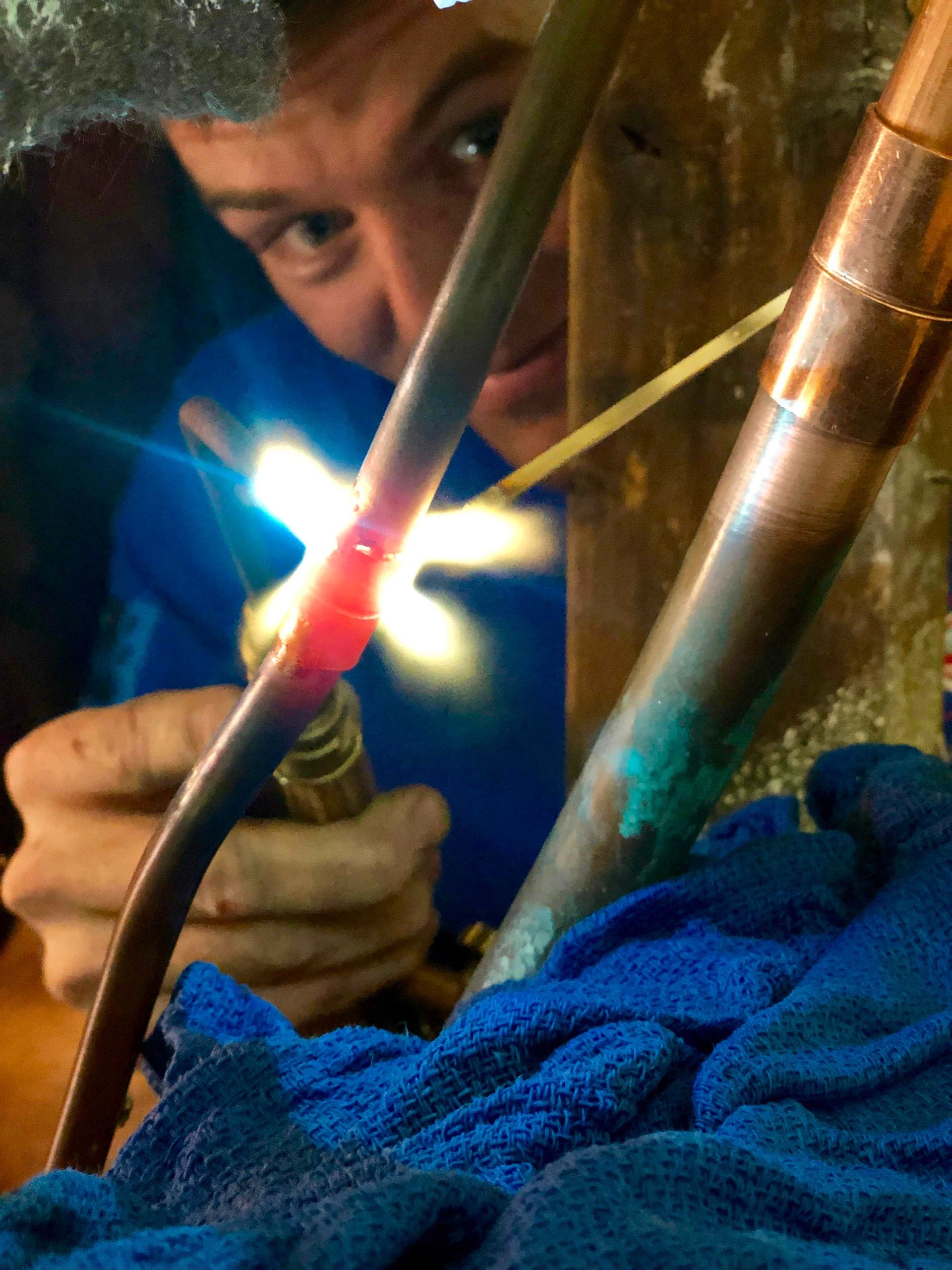 Plumber soldering copper pipes with a torch, surrounded by blue insulation in a cramped space
