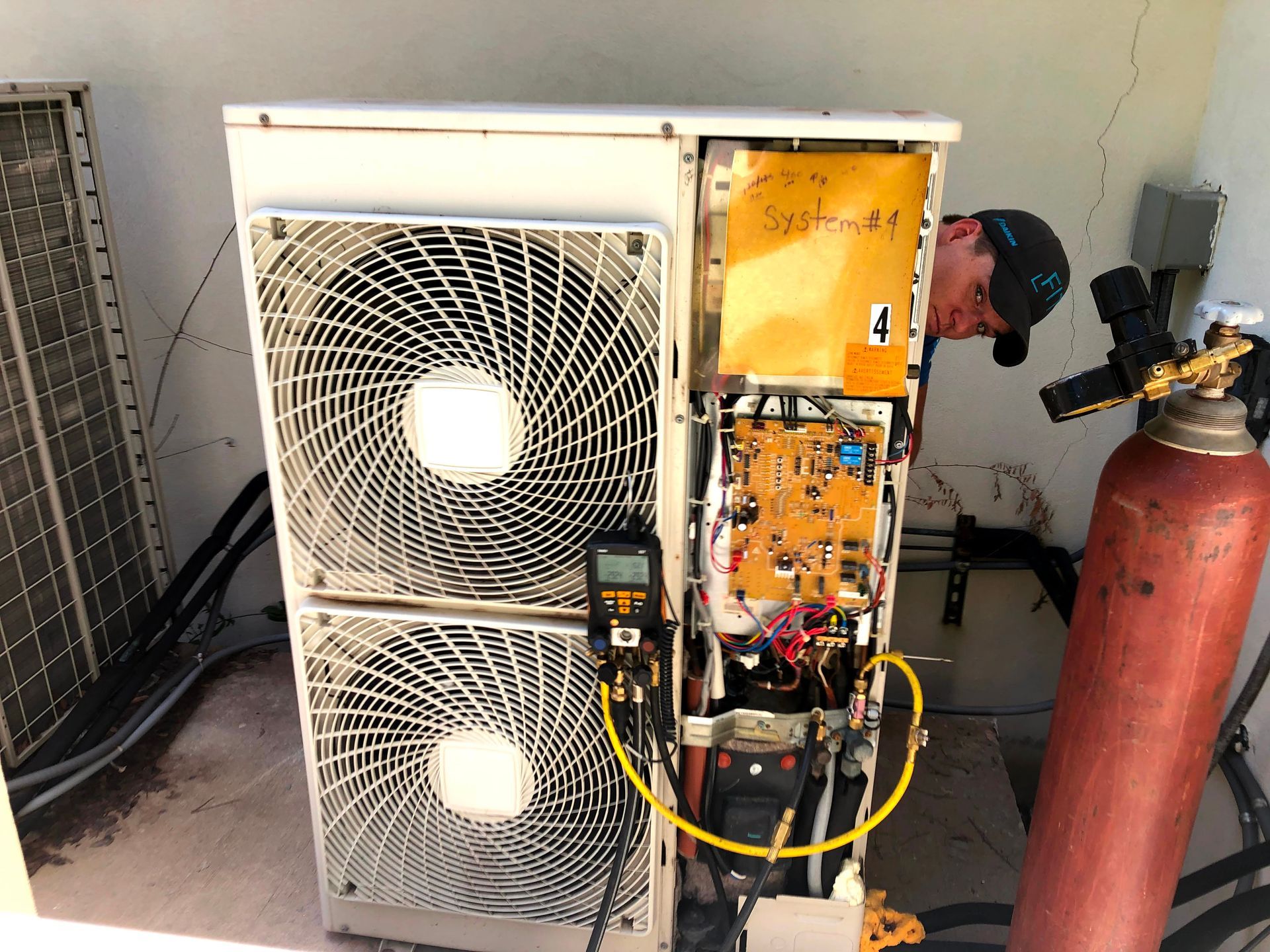 Technician repairing a heat pump with exposed wiring beside gas cylinders in a garage.