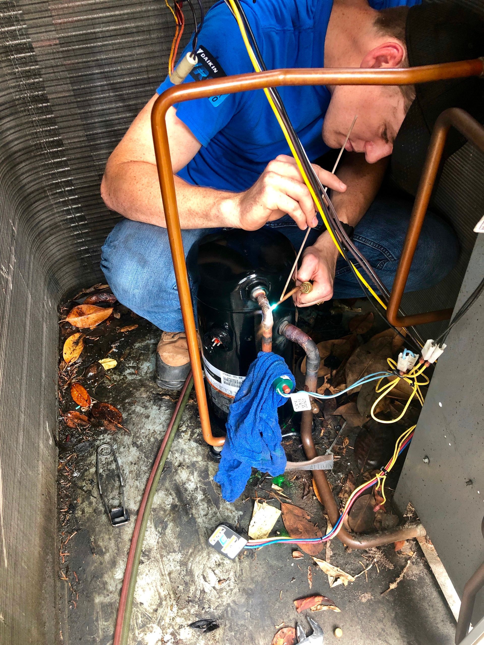 Mechanic crouches in a cramped space, working on exposed pipes with tools and wires.