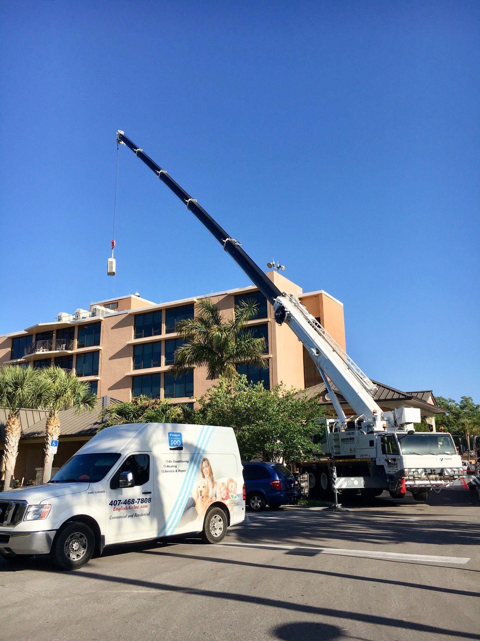 Crane lifting debris beside a tan apartment building under a clear blue sky, with work vans parked below