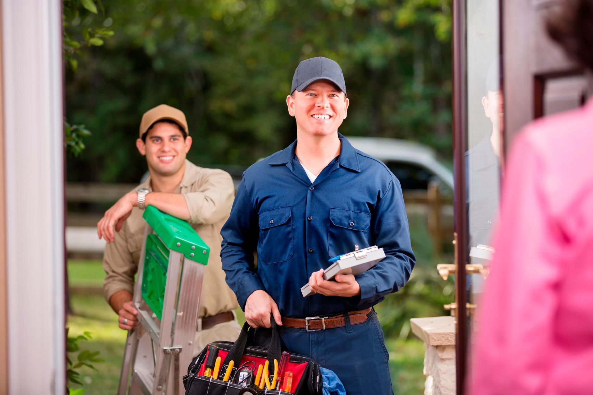 Two men smiling while carrying groceries into a home, one holding a black bag and the other a box.