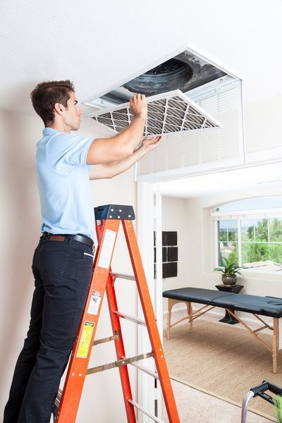 Technician cleaning a ceiling air vent from a ladder in a bright room