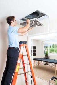 Technician cleaning a ceiling air vent in a bright room using an orange ladder