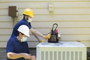 Two HVAC technicians servicing an outdoor air conditioner, one wearing a yellow hard hat and mask.