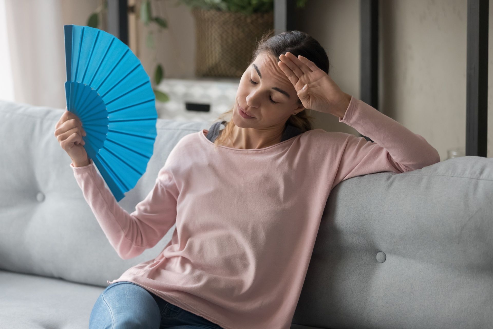 Woman on couch holding a blue fan and touching her forehead, looking overheated indoors.
