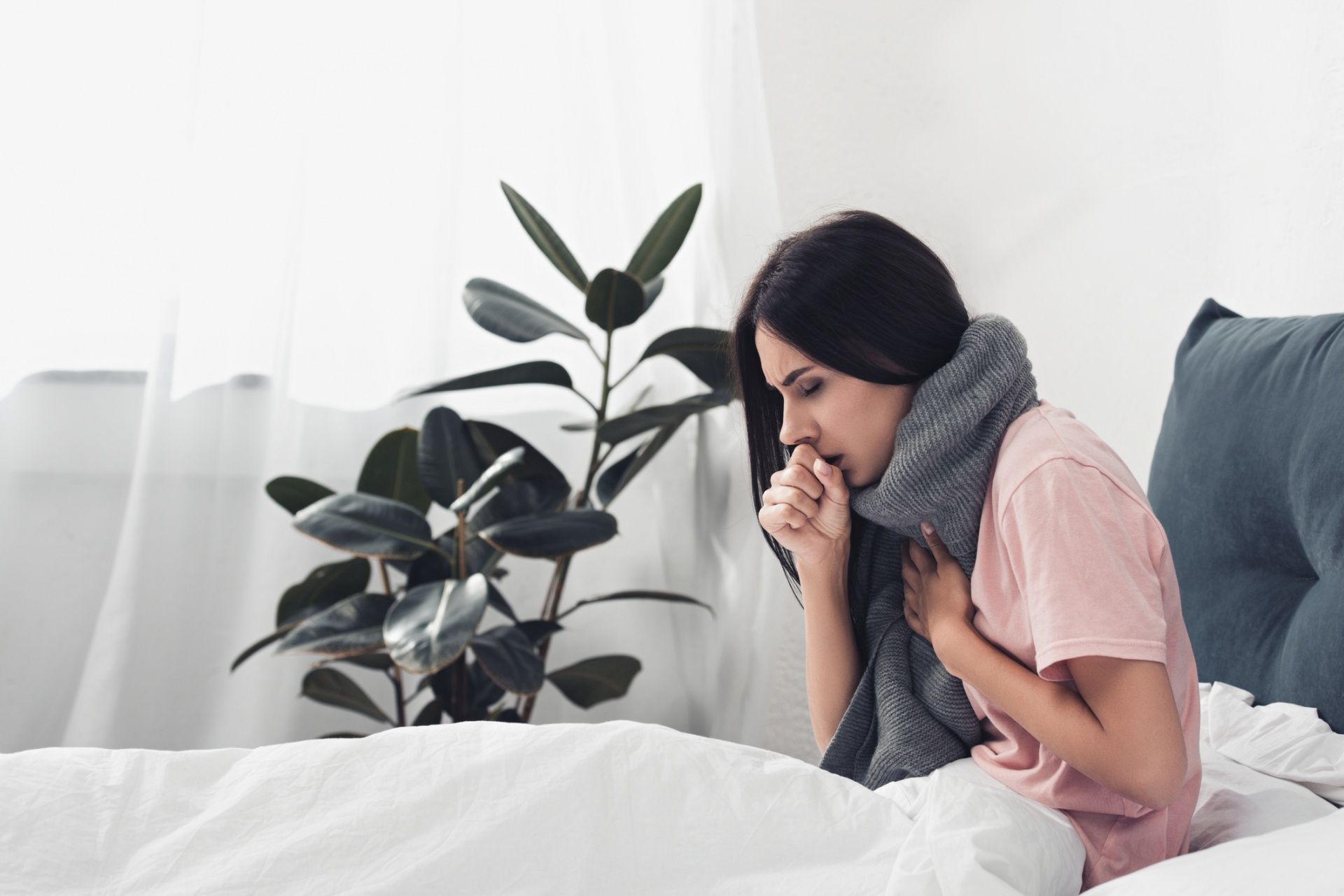 Woman sitting on a bed, coughing into her hand in a bright bedroom