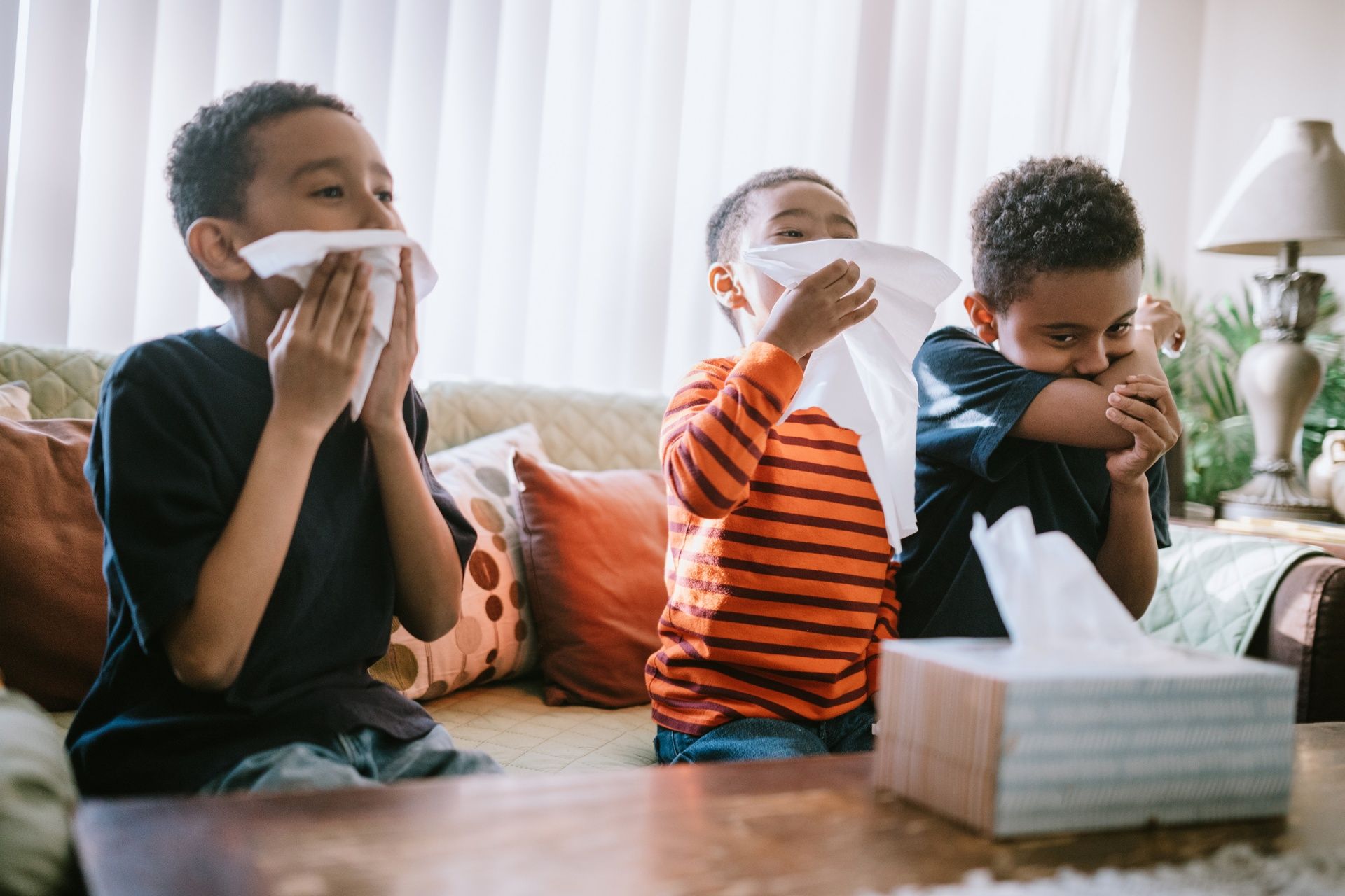 Three children on a couch blowing their noses with tissues during a cold or allergy season