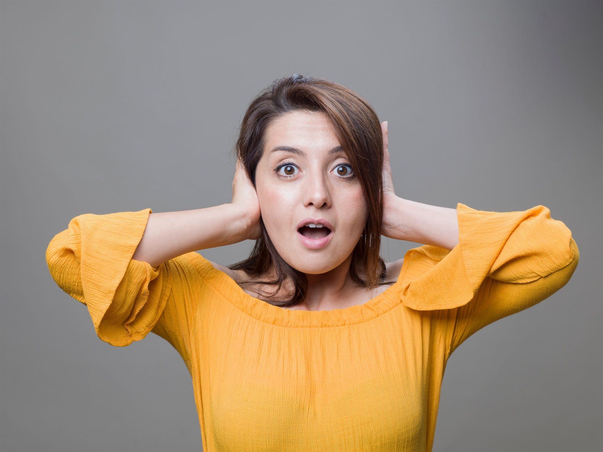 Woman in a yellow sweater covering her ears with a shocked expression against a gray background
