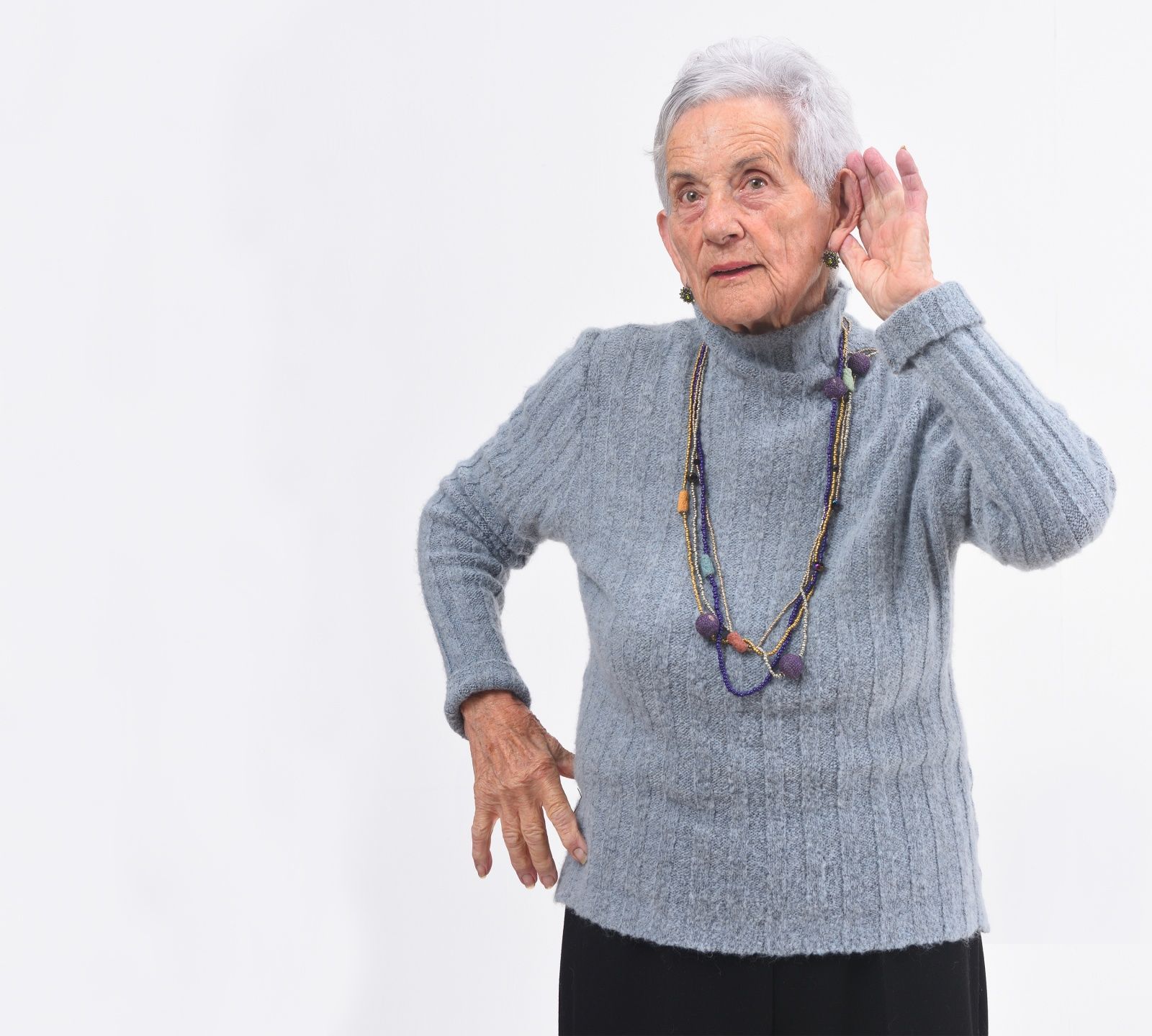 Elderly woman in gray sweater cupping her ear, standing against a white background