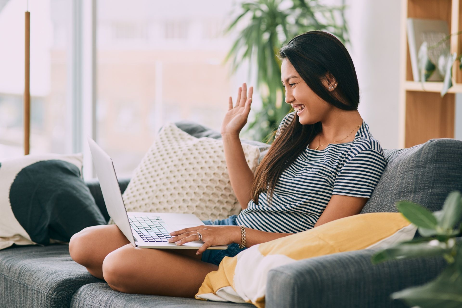 Woman sitting on a couch with a laptop, smiling and waving in a bright living room