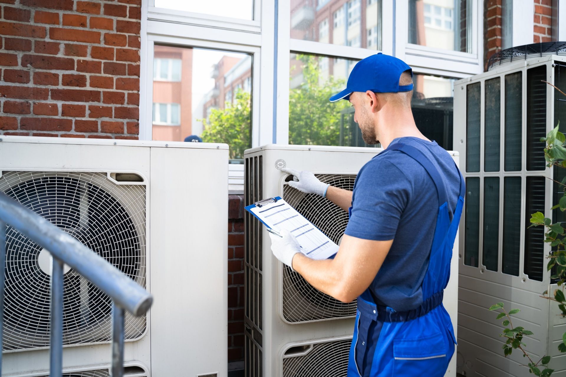 Technician in blue uniform checking outdoor air conditioning units with a clipboard by a brick building