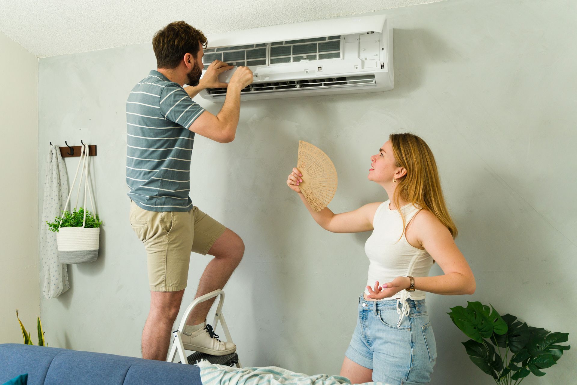 Young couple inspecting failing home air conditioning unit while cooling off indoors.