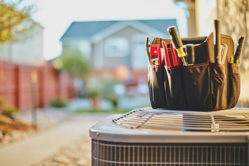 Tool bag on an outdoor HVAC unit in a suburban yard, with house and fence blurred in the background