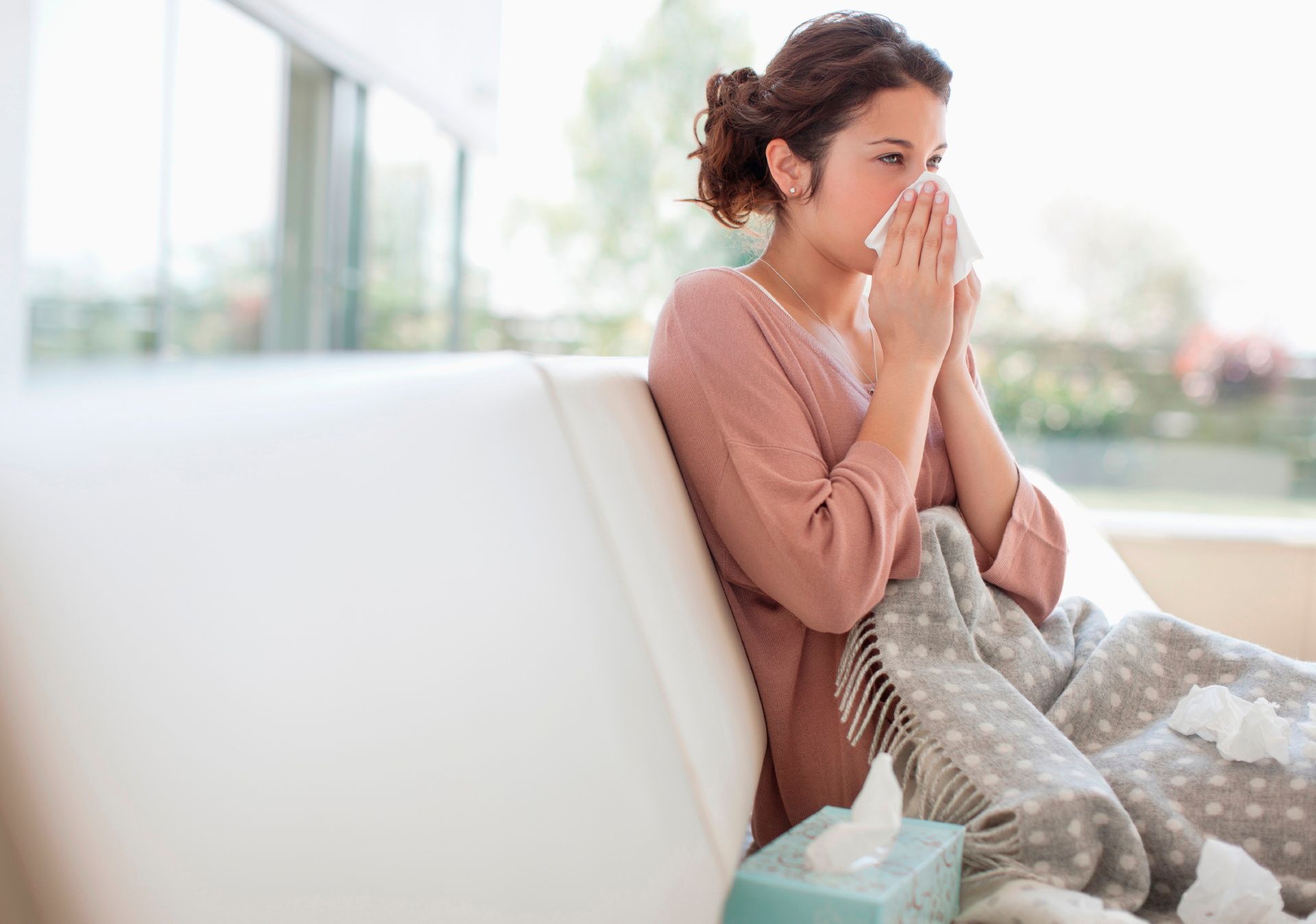 Woman on couch holding tissue to her nose, looking unwell in a bright living room.