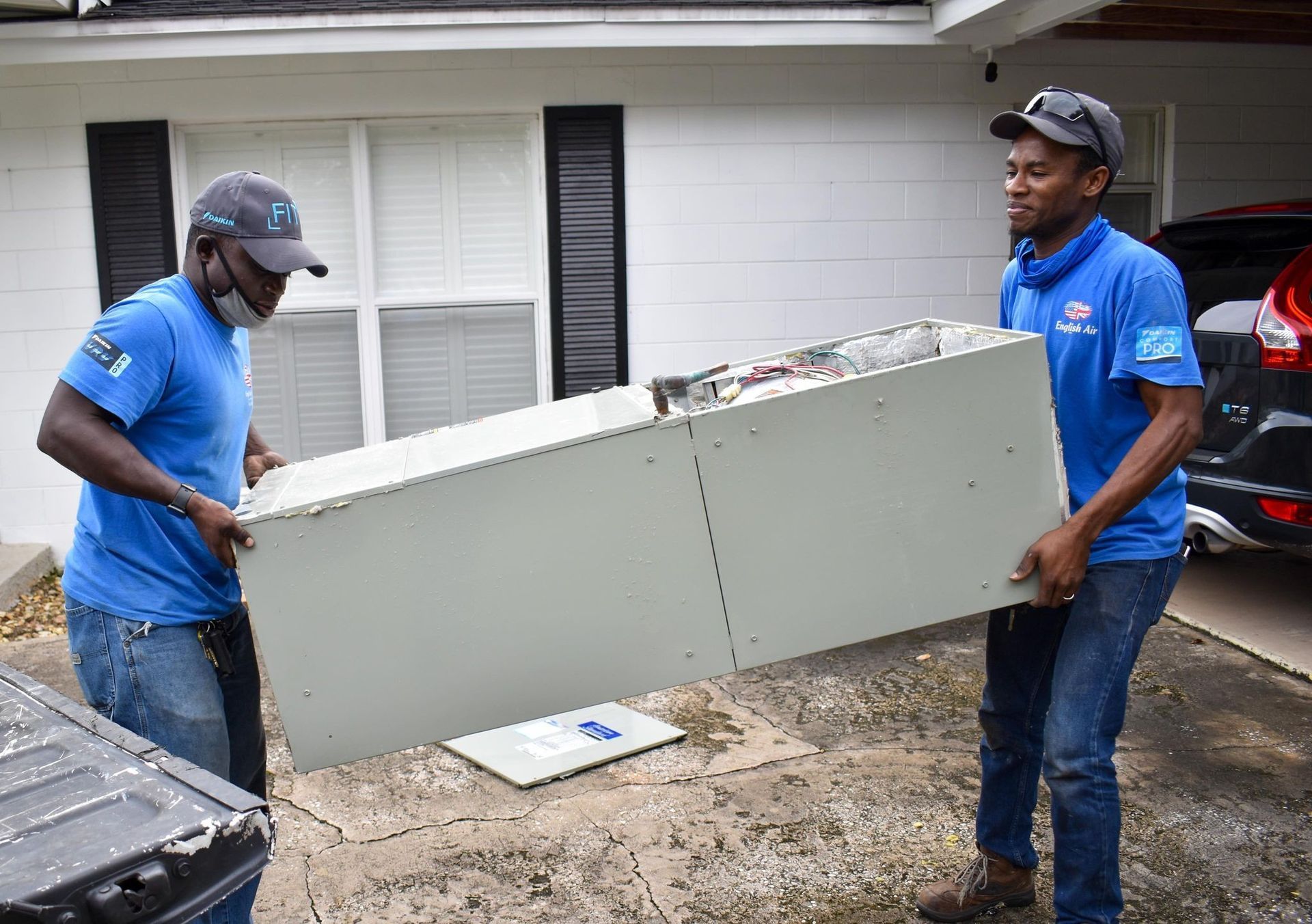 Two workers in blue shirts carry a large white cabinet outside a house.