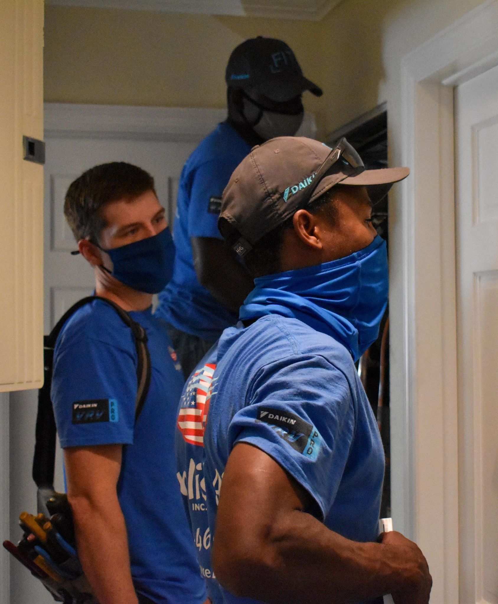 Three men in blue shirts and masks standing in a hallway, looking into a doorway.