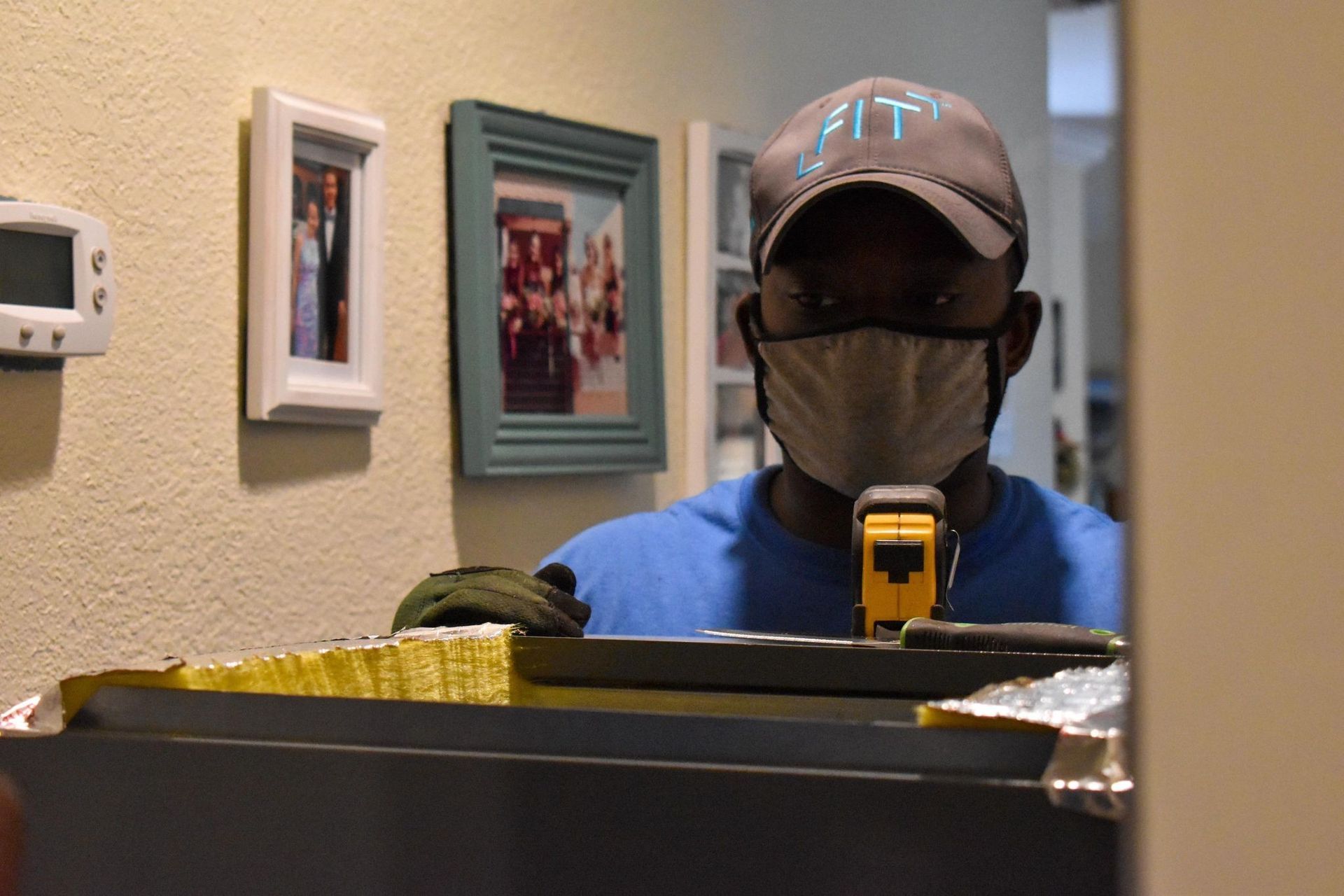 Masked person in a blue shirt and cap standing behind a yellow-and-black barrier indoors