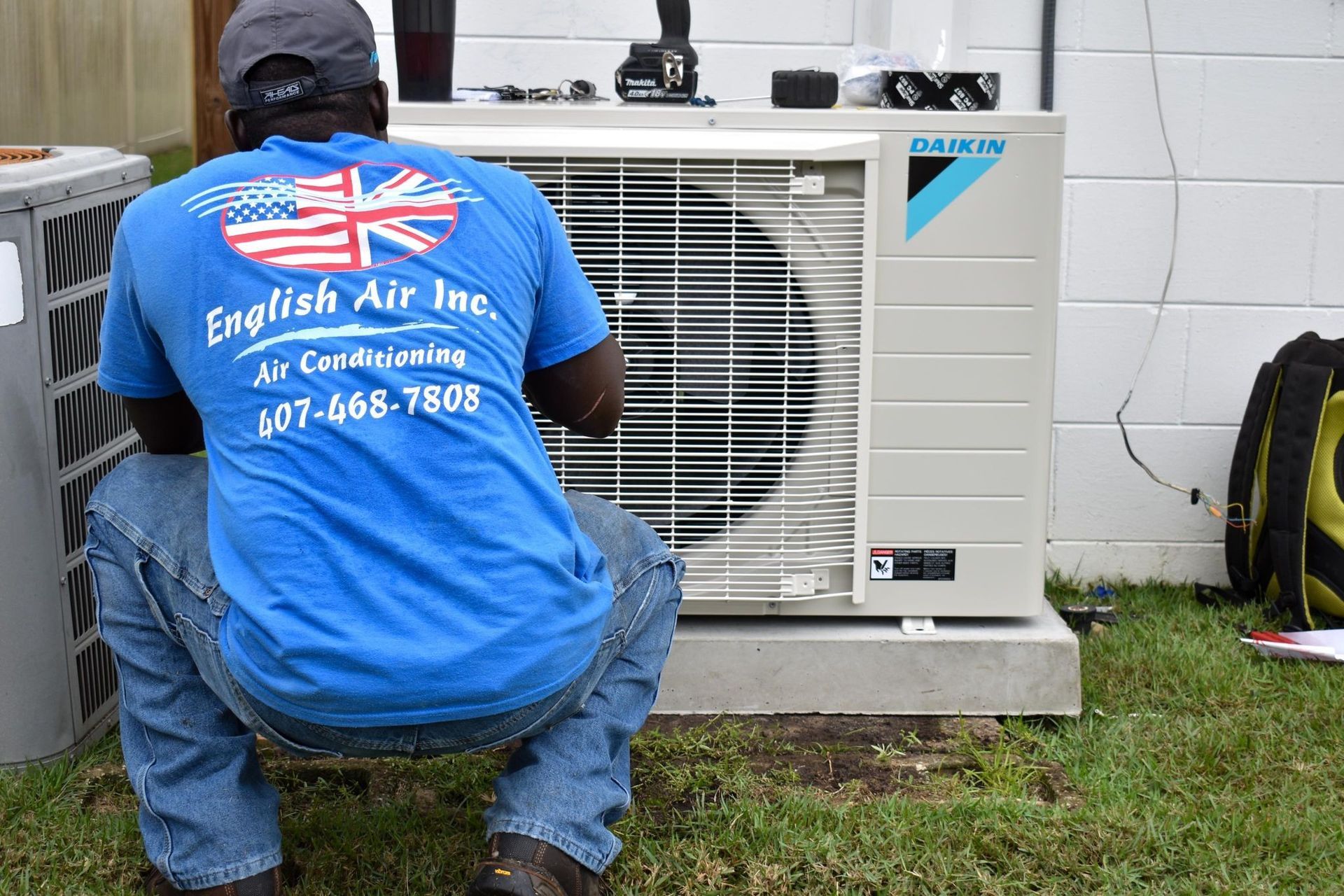 Technician in blue shirt kneels beside an outdoor air conditioning unit, inspecting it on a grassy area.
