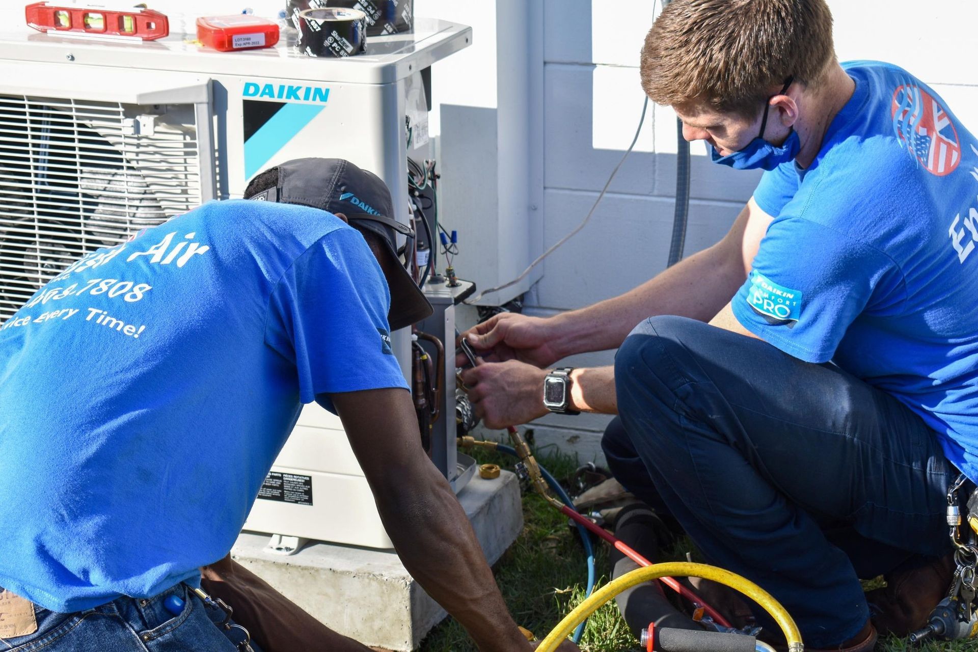 Two technicians in blue uniforms repair an outdoor Daikin air conditioner unit.