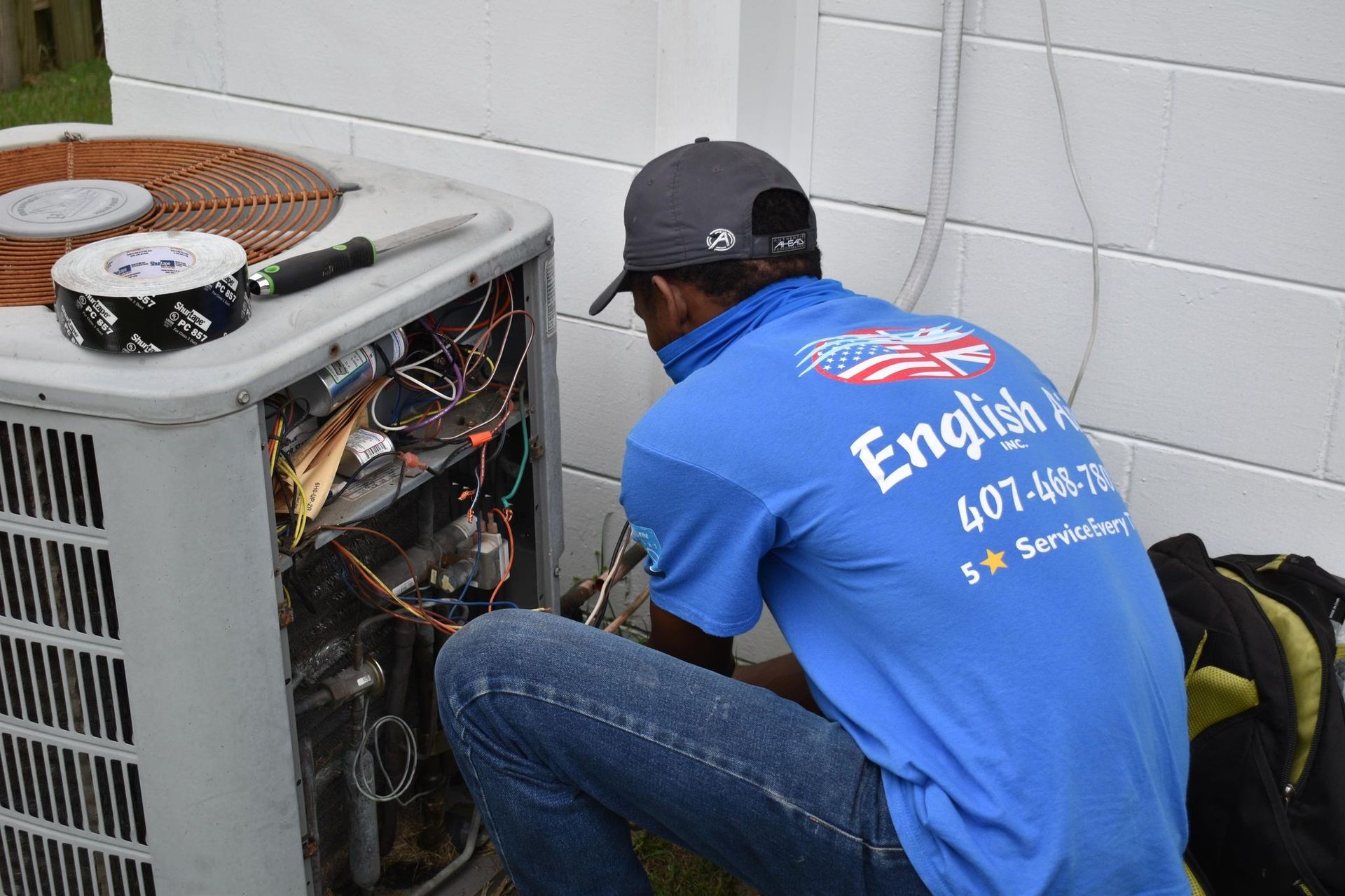 Technician in blue shirt repairs an outdoor air conditioner unit beside a wall.