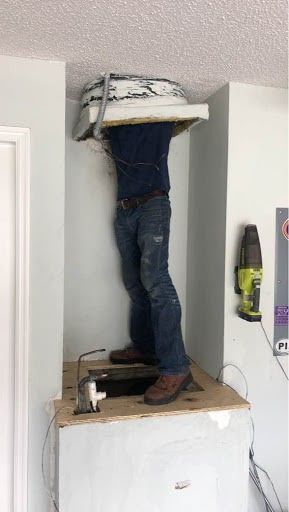 Person standing on a cabinet, reaching up inside an open wall space with a white appliance or unit above.