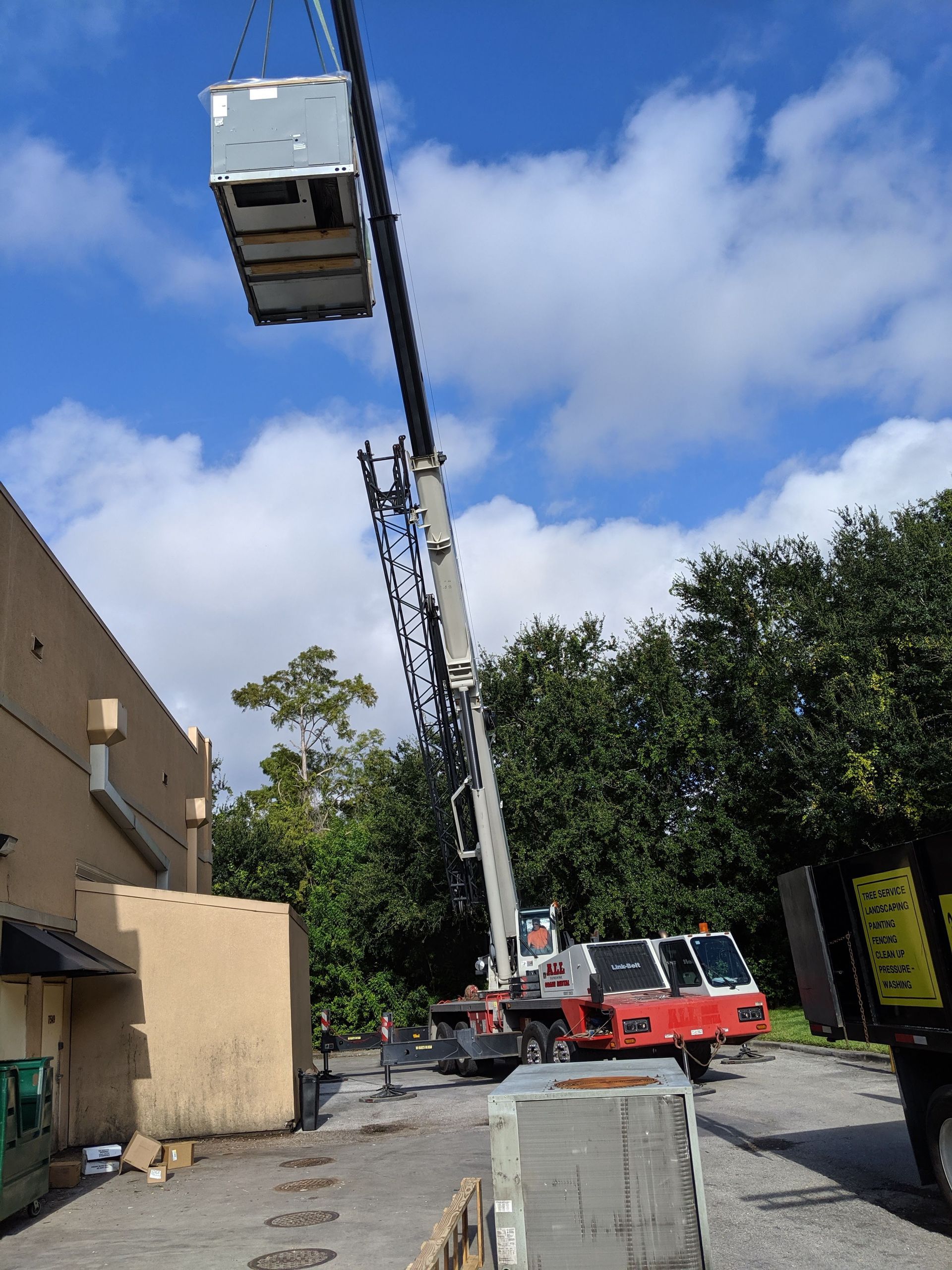 Crane lifting a white shipping container above a worksite beside a building and trees under a blue sky.