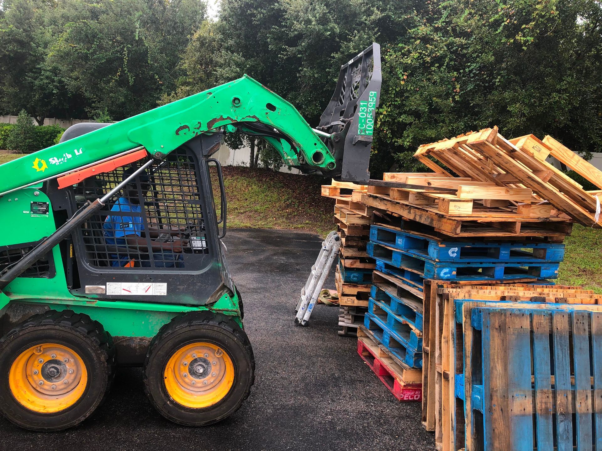 Green skid-steer loader lifting wooden pallets beside a stack of blue and red pallets outdoors