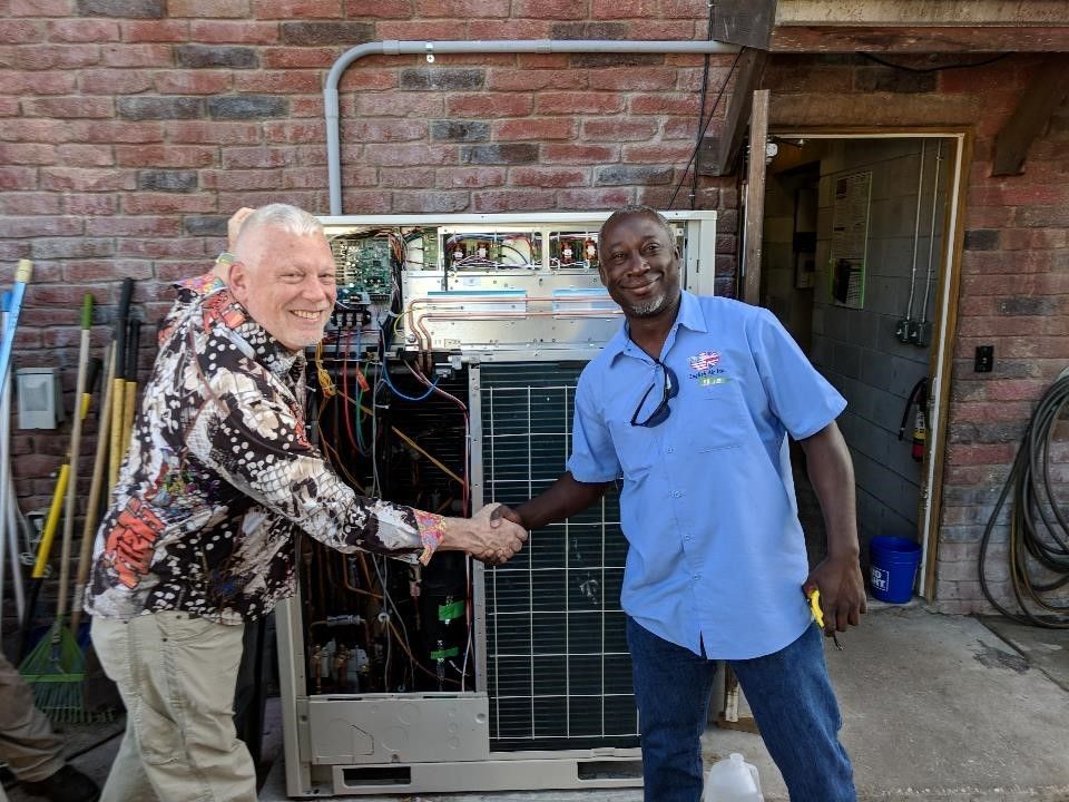 Two men shake hands beside a large HVAC unit in front of a brick building.