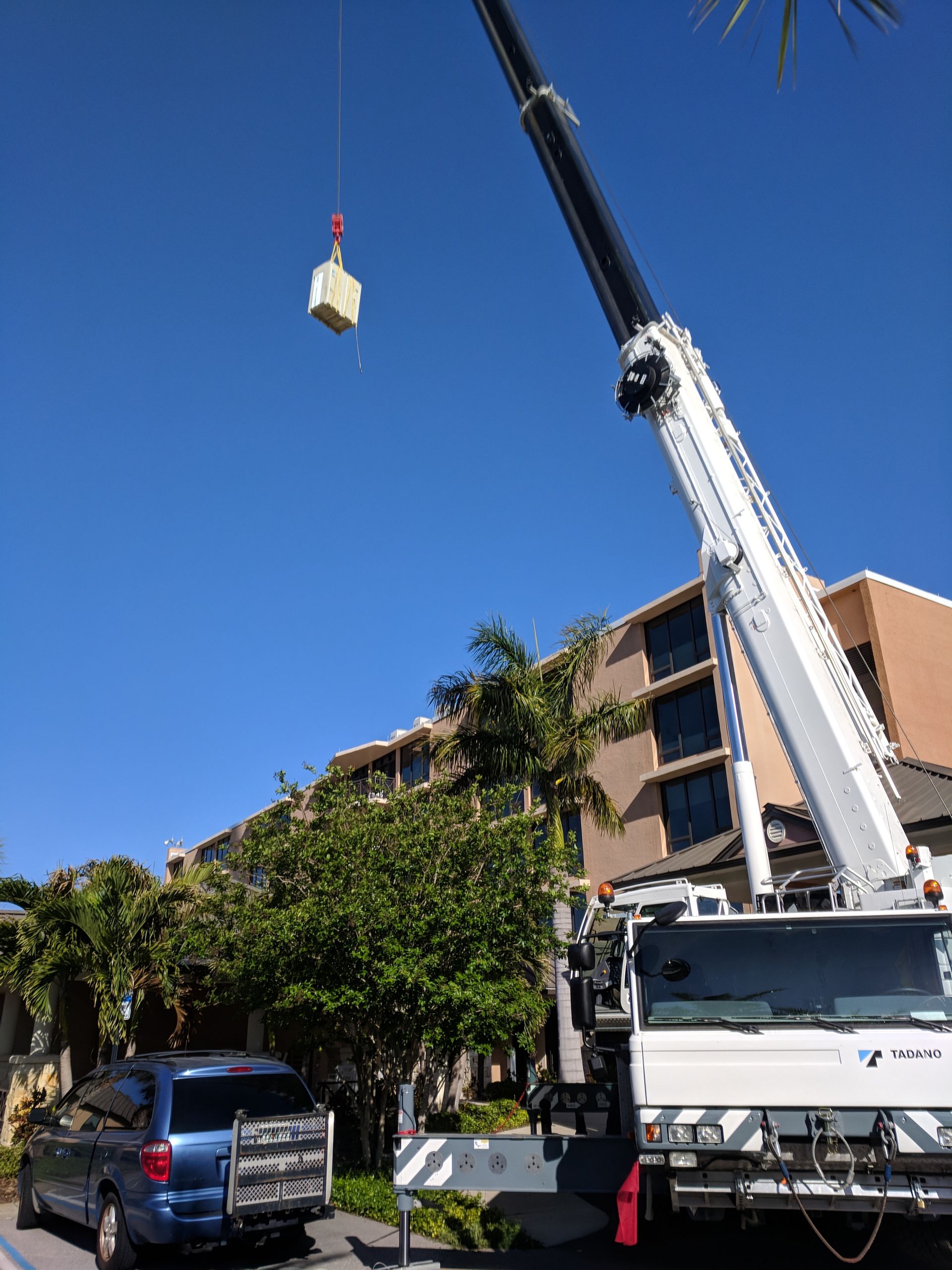Crane lifting a small white box beside a blue car and apartment building under a clear sky