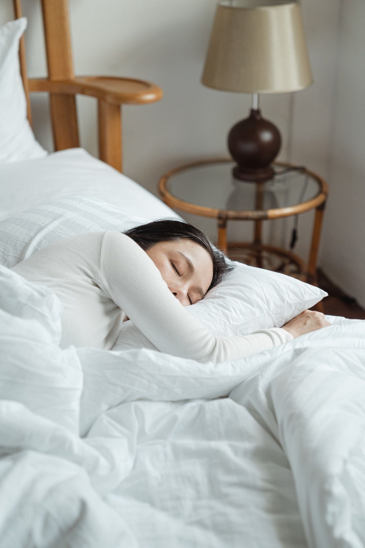 Person sleeping in a white bed beside a wooden chair and bedside lamp.