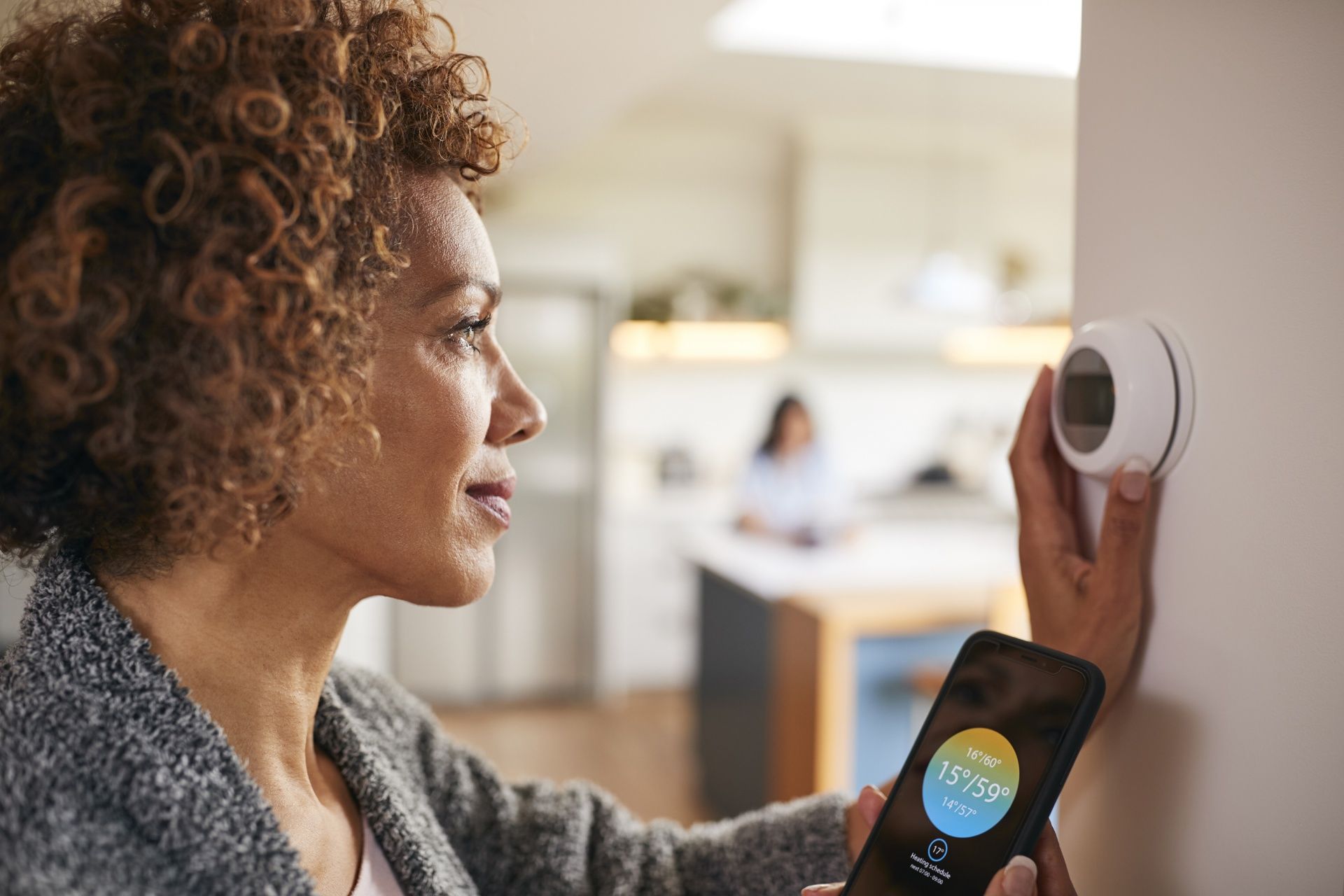 Person adjusting a wall thermostat while holding a smartphone with a temperature app open