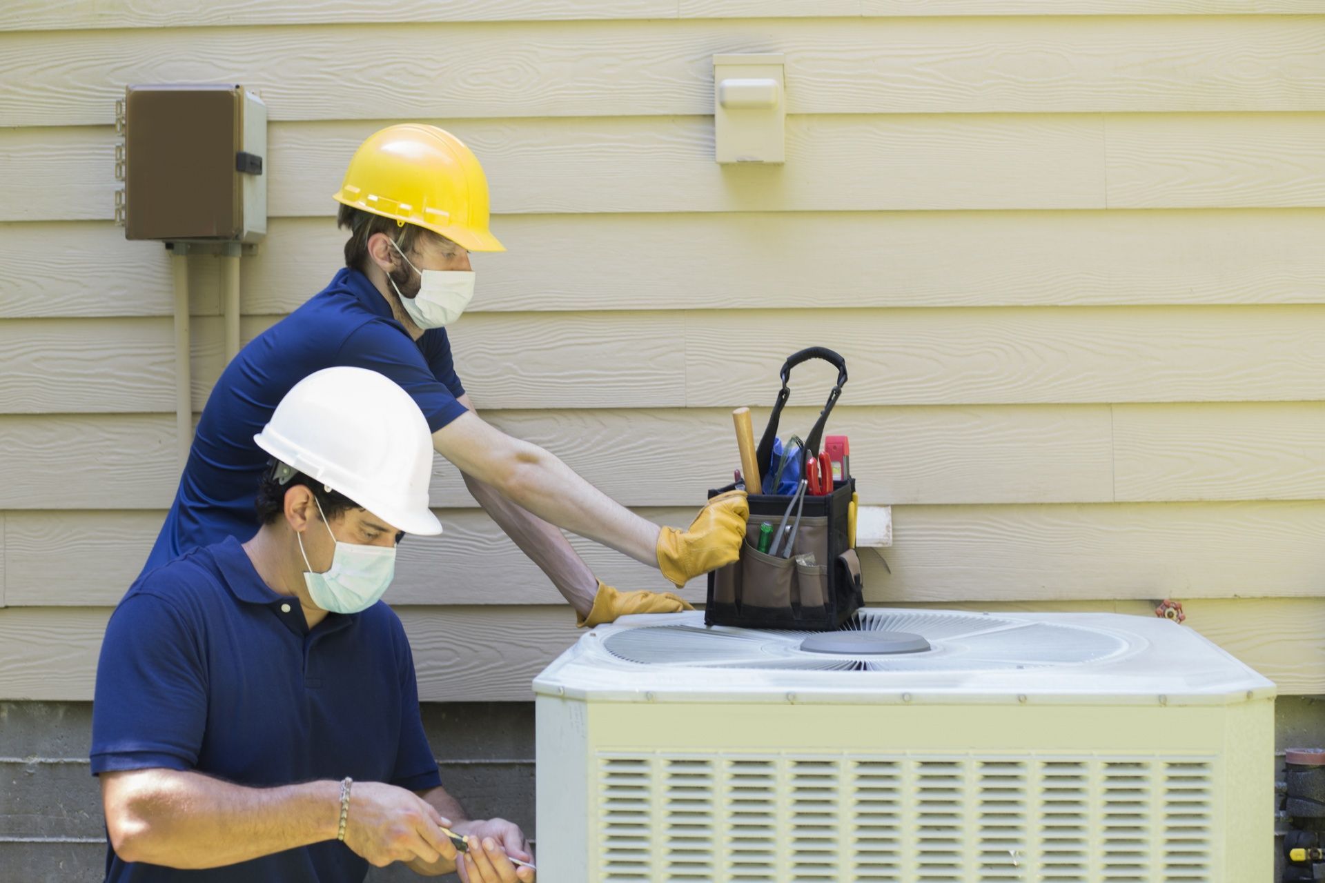 Two workers in hard hats servicing an outdoor air conditioner unit beside a house