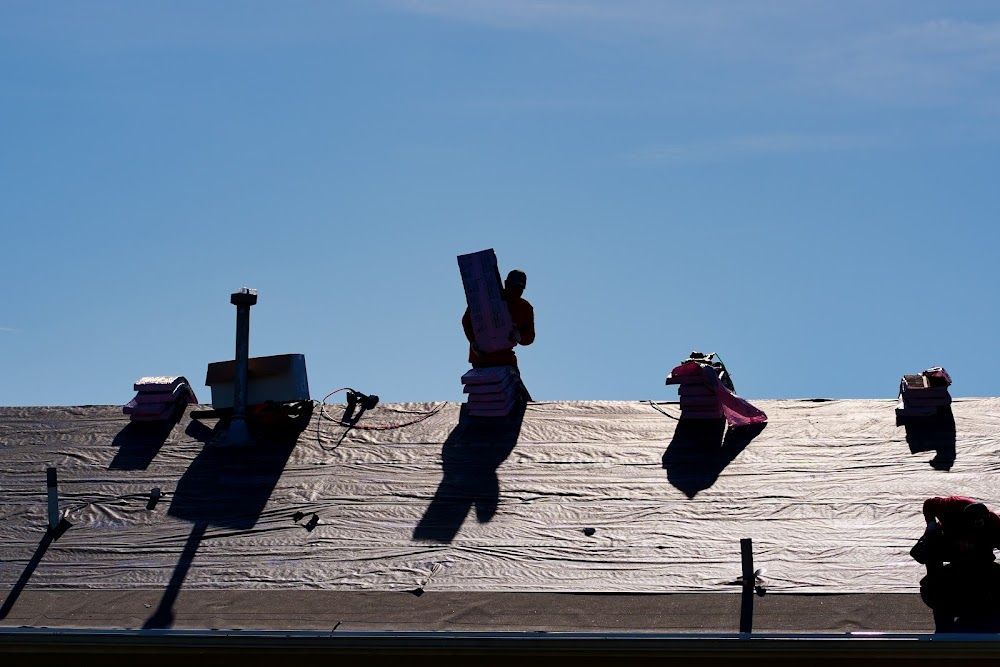 Roofer carrying material on a dark roof with a clear blue sky background.